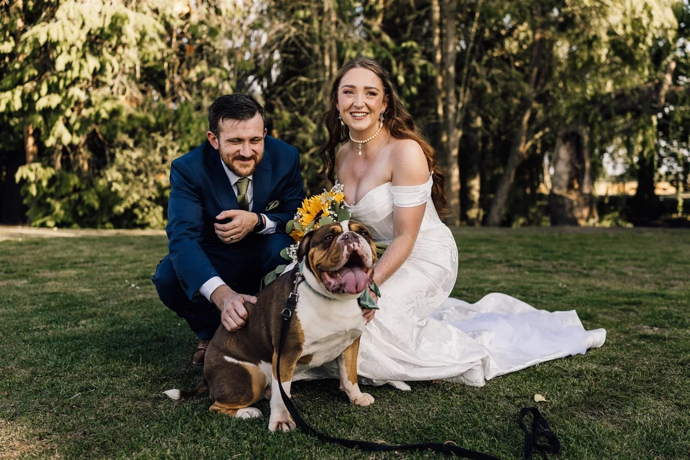 A bride and groom are kneeling on grass outdoors, smiling at the camera, with a large happy bulldog sitting in front of them. The bride is wearing a white off-shoulder wedding gown and pearl jewelry, and holding a bouquet. The groom is dressed in a n