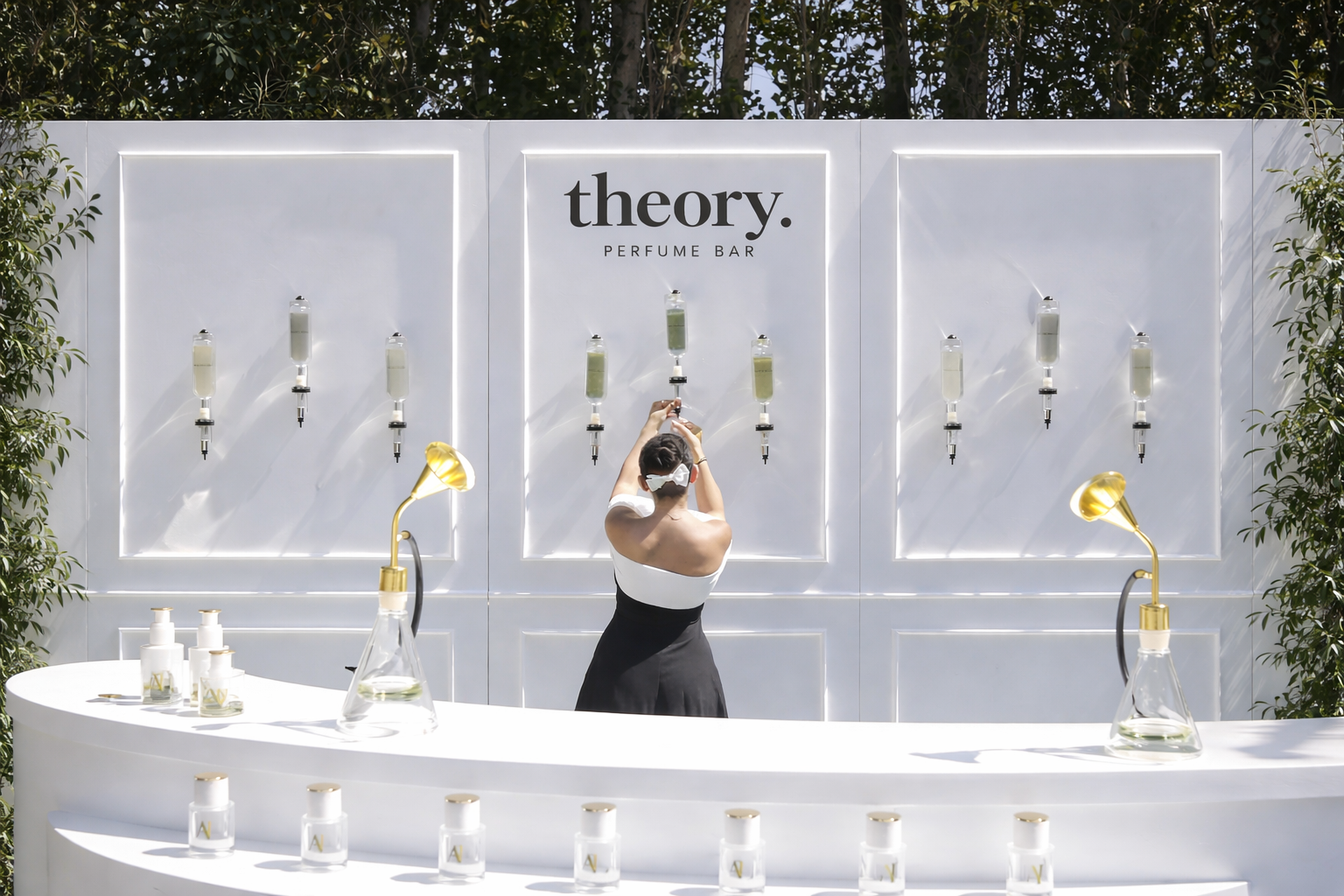 A person with short dark hair and a white dress with black trim stands at a perfume bar labeled 'theory PERFUME BAR,' pouring perfume from a glass container onto a test strip. The bar has white bottles and gold lamps, set outdoors with greenery in the background.