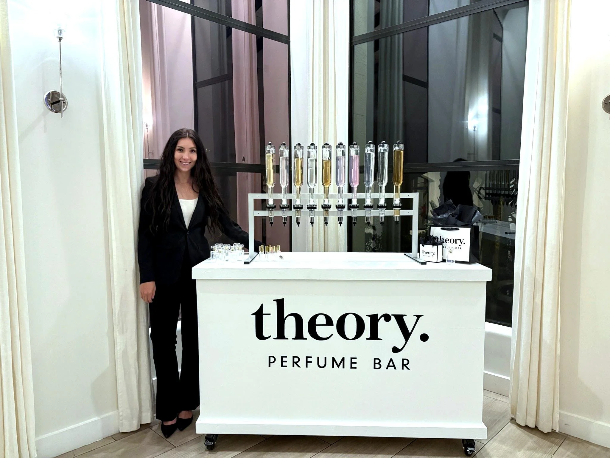 Woman standing next to a white perfume bar with the word 'theory' in bold black letters, bottles of perfume with spray tops on display.