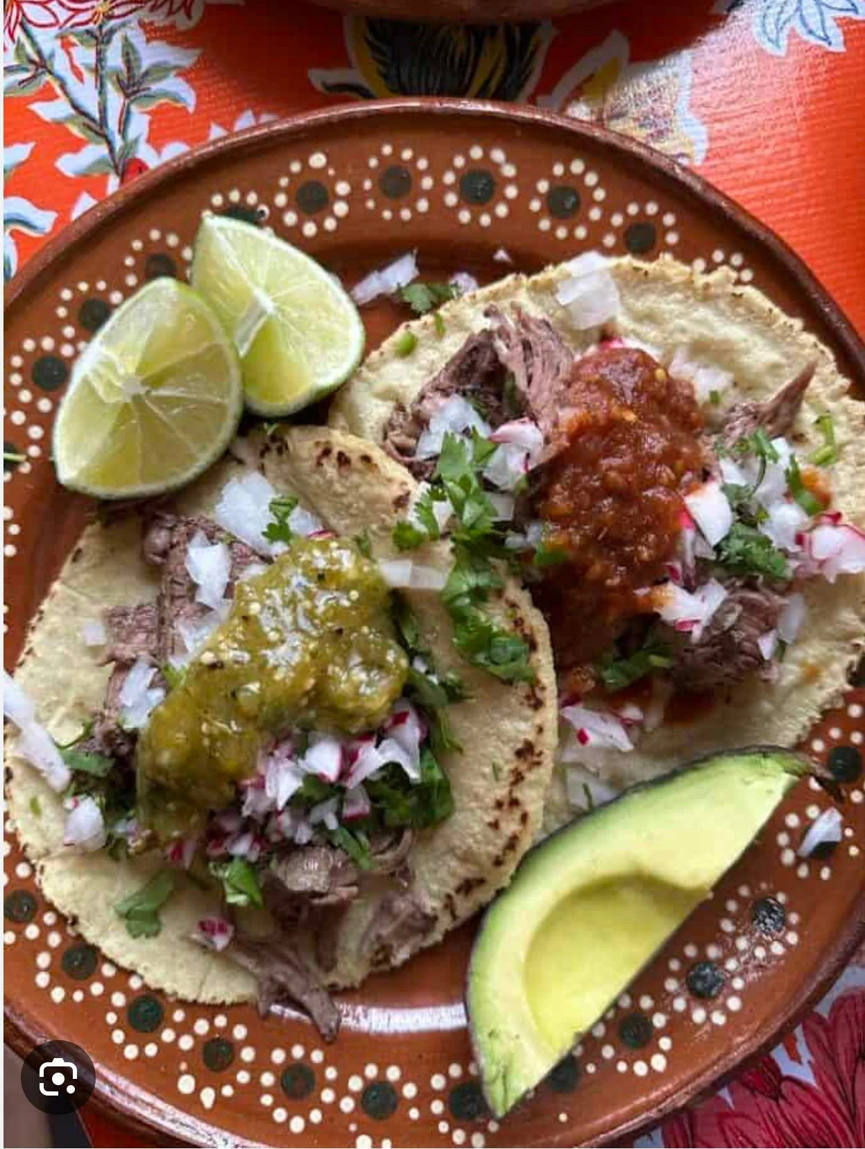 Three tacos on a decorative orange plate with lime wedges, topped with diced onions, cilantro, and various salsas.