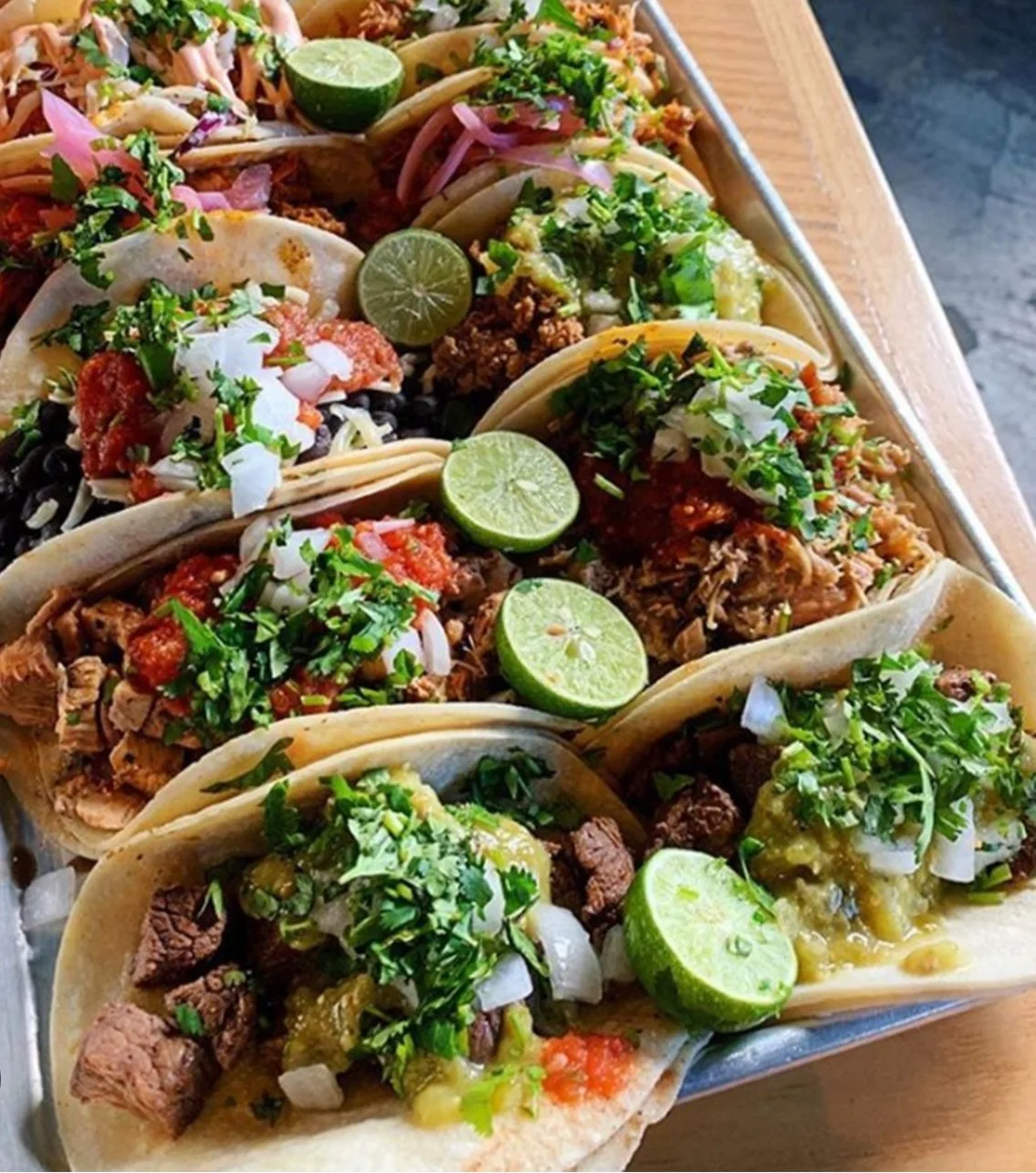 Assorted tacos topped with chopped cilantro and onions, lime wedges on a tray.
