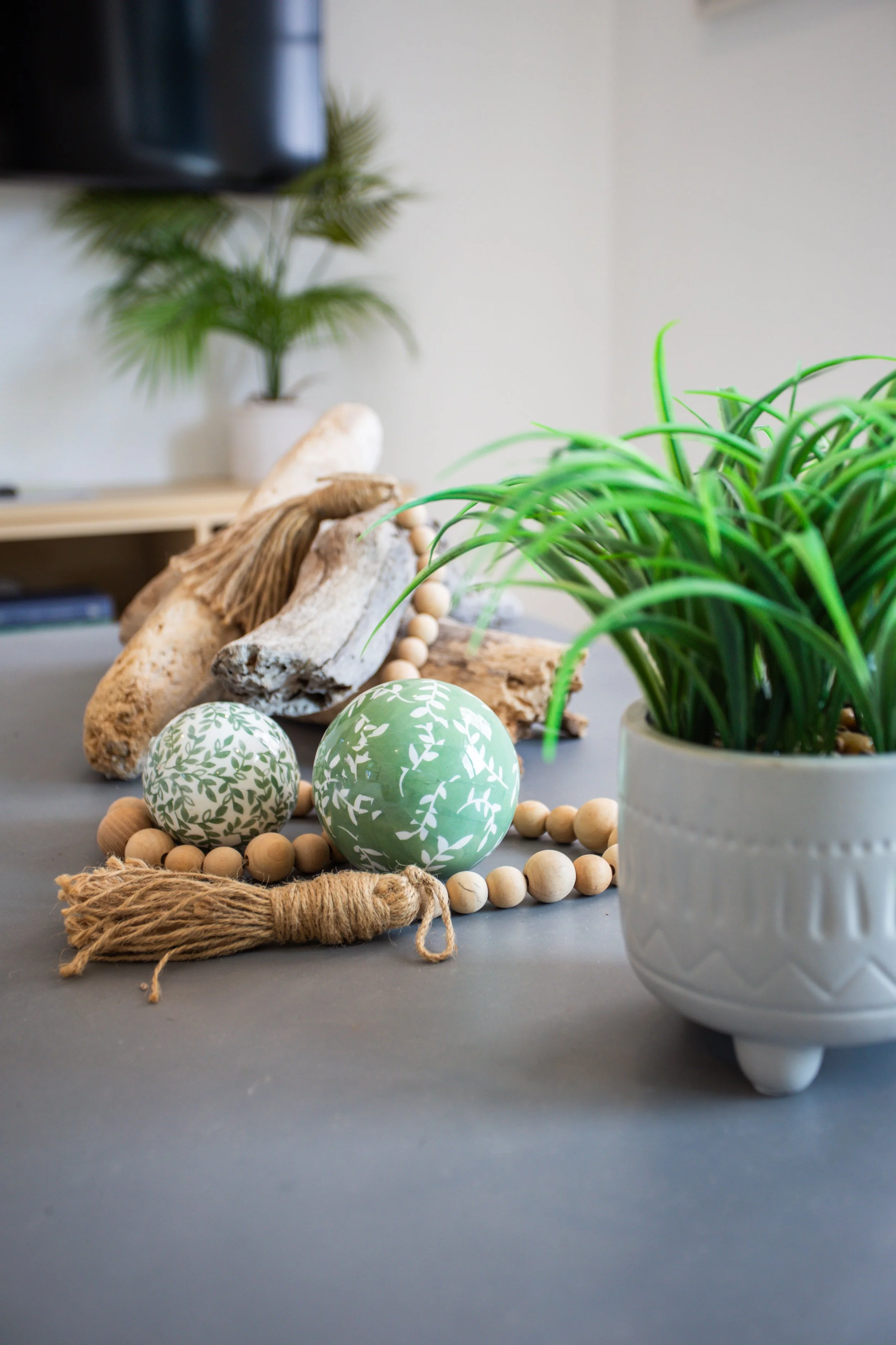 Decorative indoor arrangement featuring green patterned balls, wooden bead garland, driftwood, and potted green plant on a gray surface.