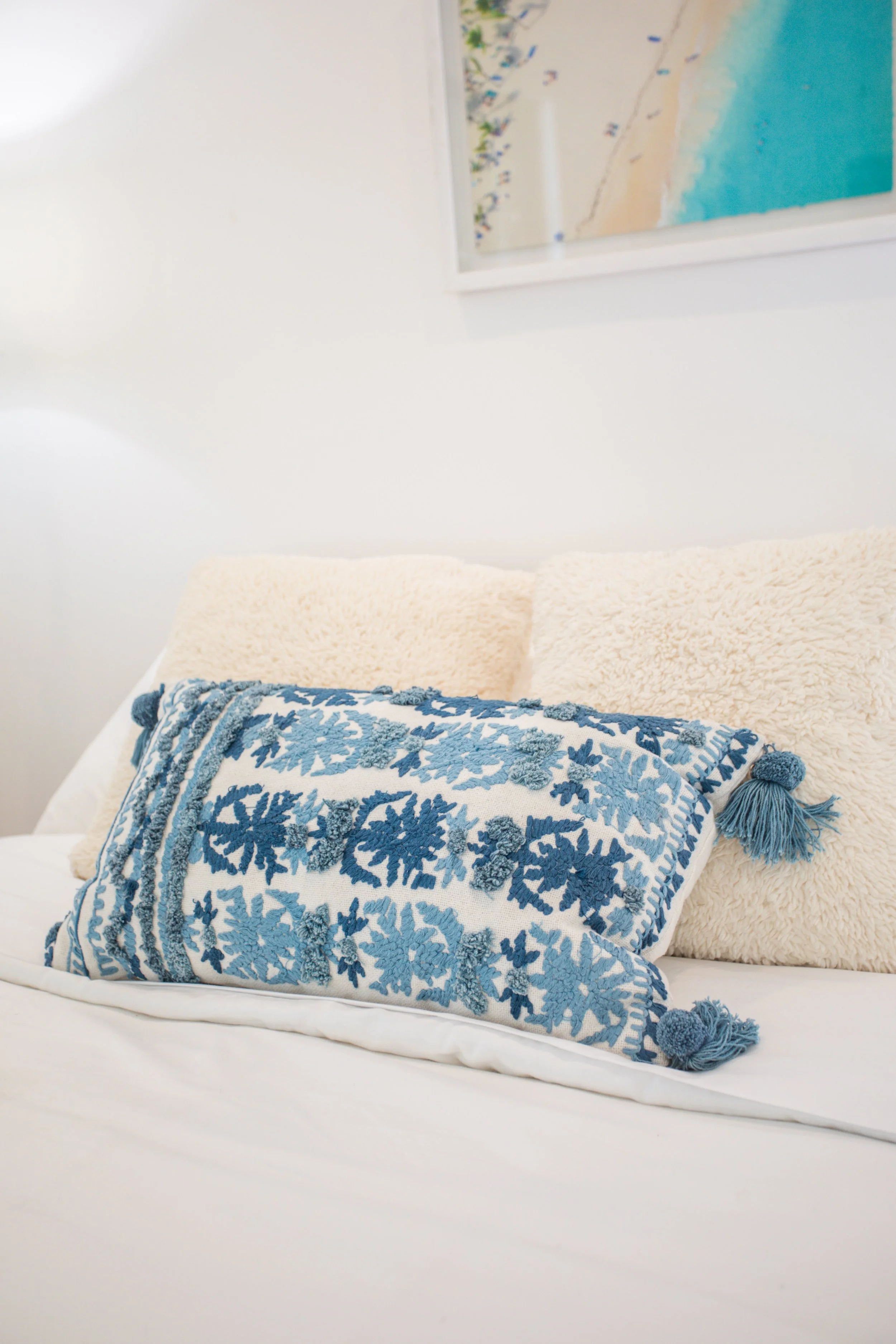 A cozy bedroom with a bed featuring white and cream pillows, and a decorative blue and white embroidered pillow with tassels, against a white wall with a beach-themed painting.