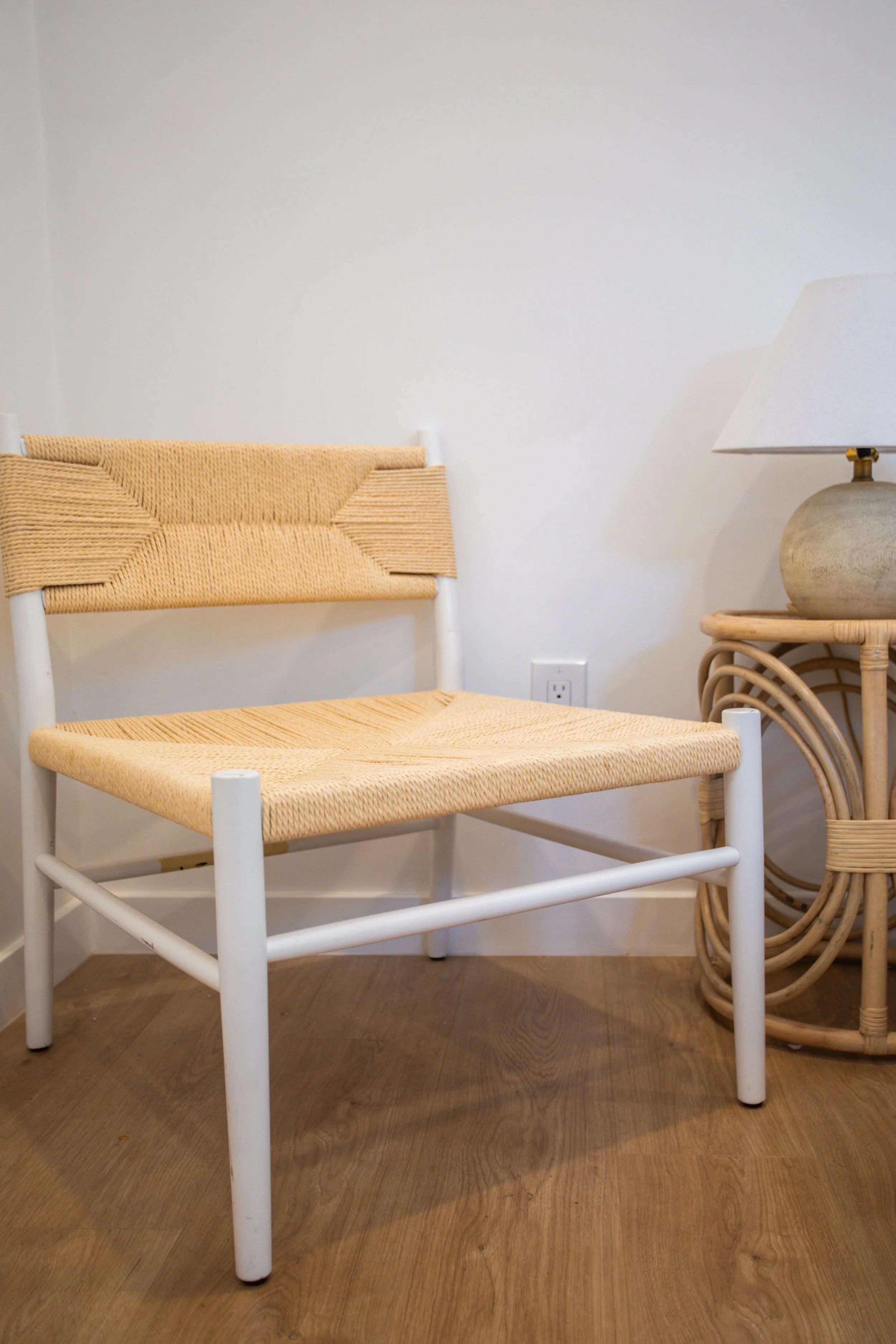 A beige woven chair with a white frame, positioned next to a rattan side table with a lamp, on a wooden floor against a plain white wall.
