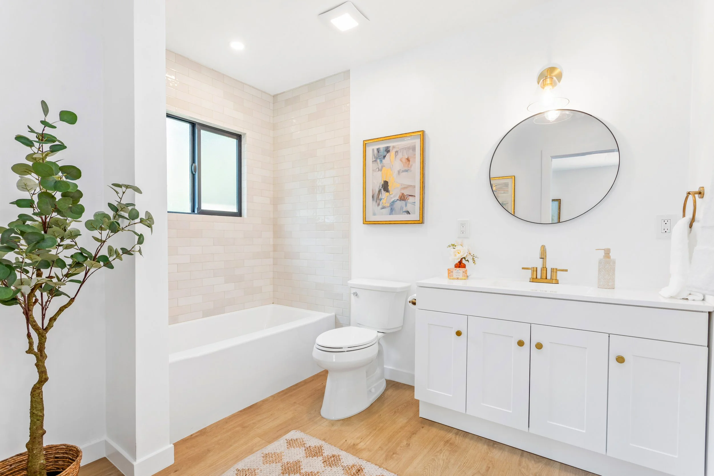 Bright bathroom with a white vanity, round mirror, gold fixtures, white bathtub with tiled wall, small window, framed artwork, potted plant, and wooden floor.
