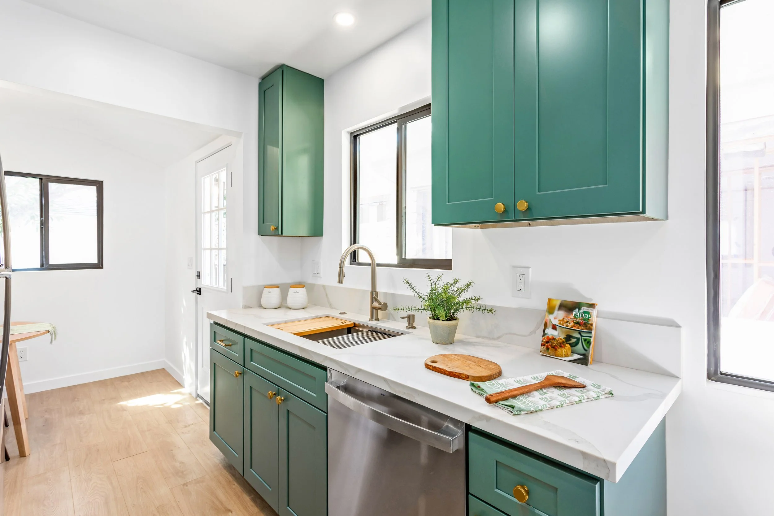 Modern kitchen with green cabinets, white marble countertops, and stainless steel dishwasher. Features a window above the sink, potted plant, chopping board, and kitchen magazines.