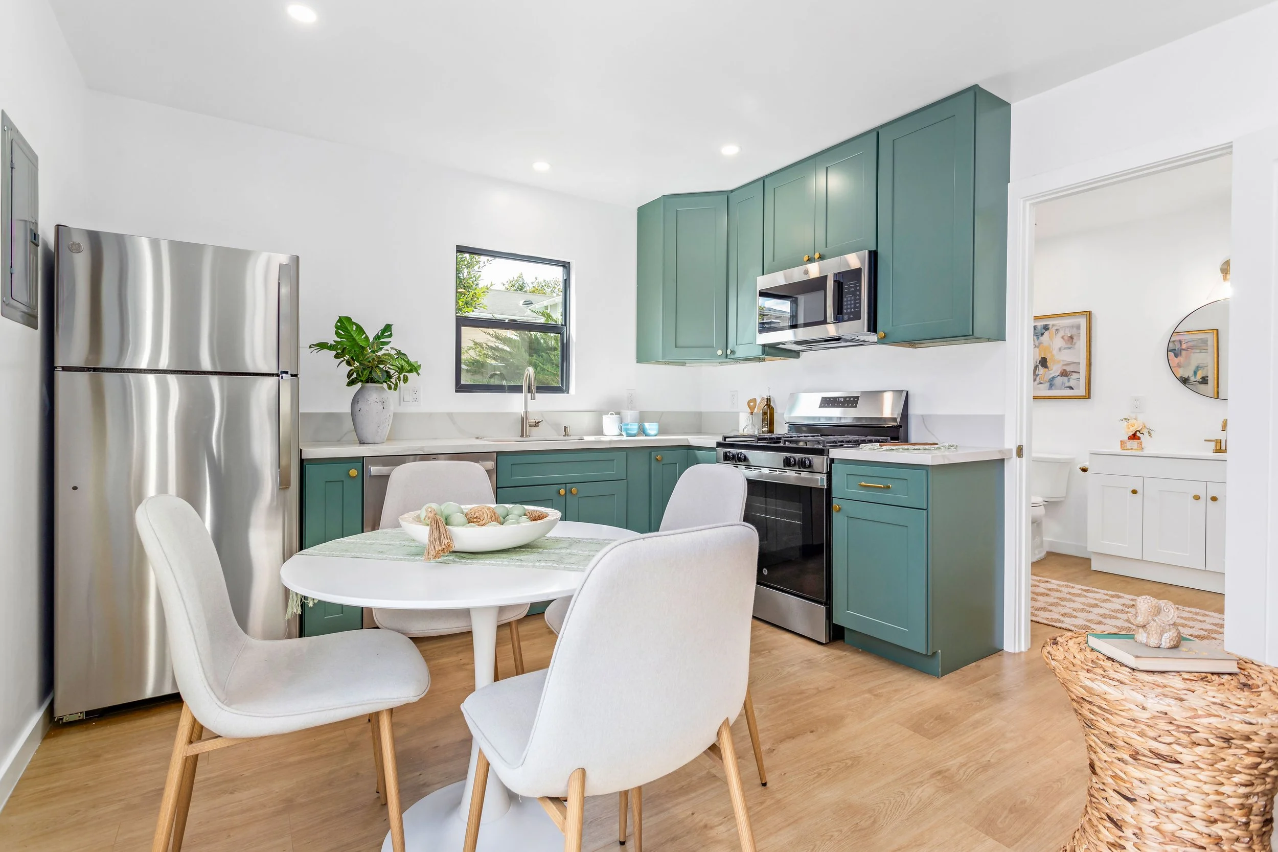 Modern kitchen with teal cabinets, stainless steel appliances, a small round table with four white chairs, a window with greenery outside, and a glimpse of a bathroom in the background.