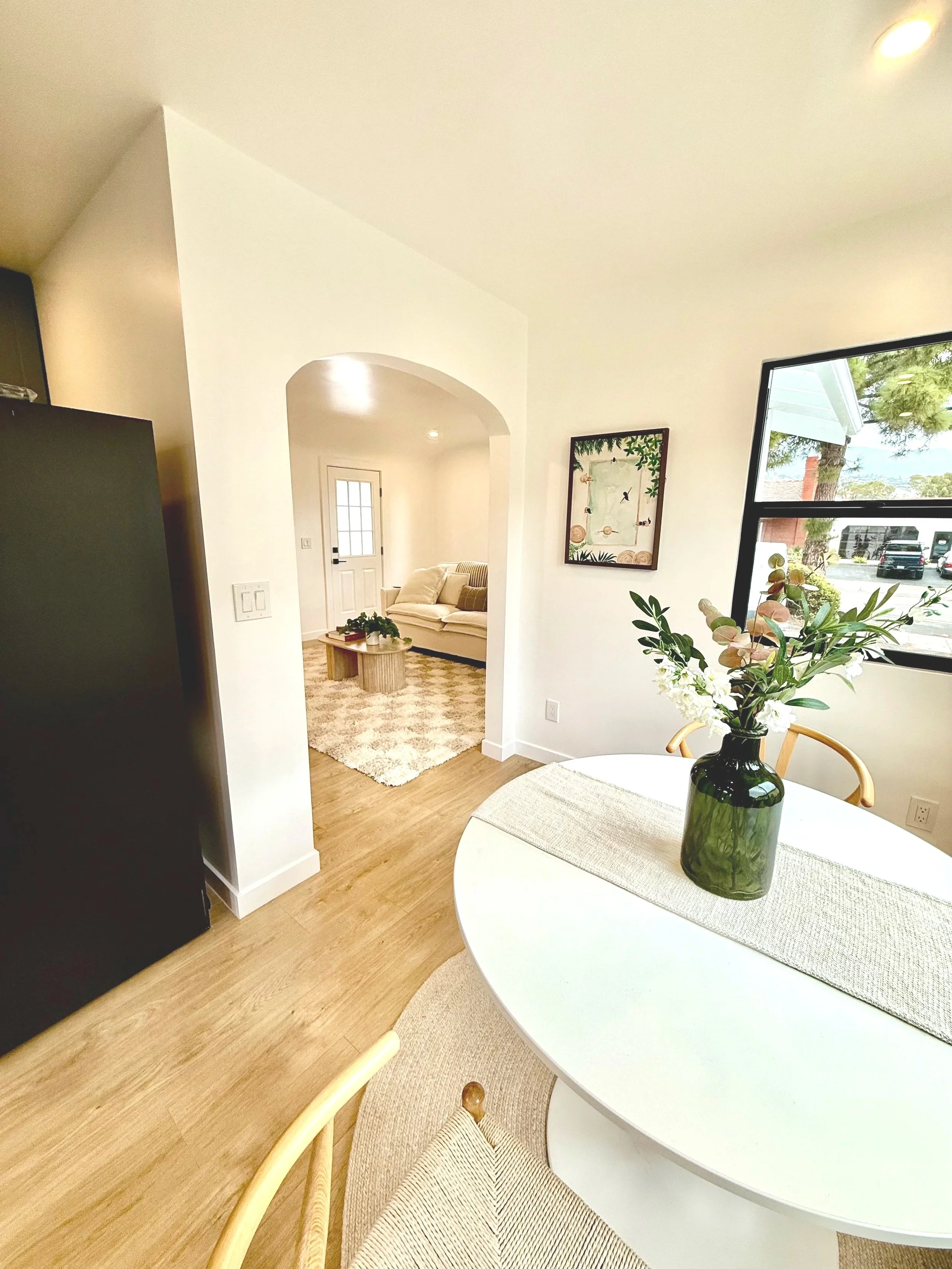 A cozy dining area with a round table, a green glass vase with white flowers, and a window showing a tree outside. In the background, a living room with a beige sofa, a white door, and a patterned rug is visible.