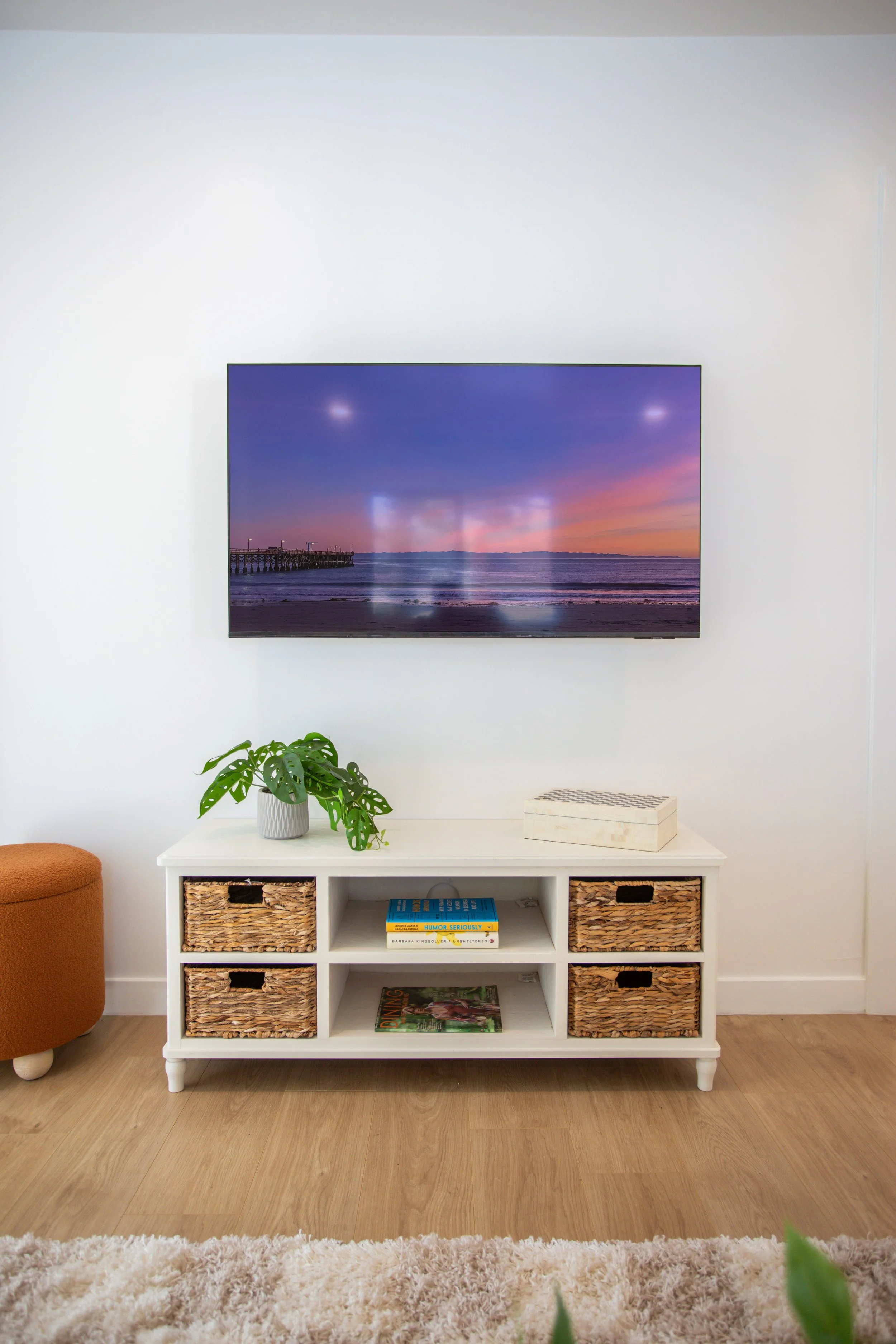 Living room with a white TV stand, framed by rack baskets, holding a potted plant and books, and a large flat-screen TV displaying a sunset over the ocean. There's a small orange ottoman on the left and a plush beige rug on the wooden floor.