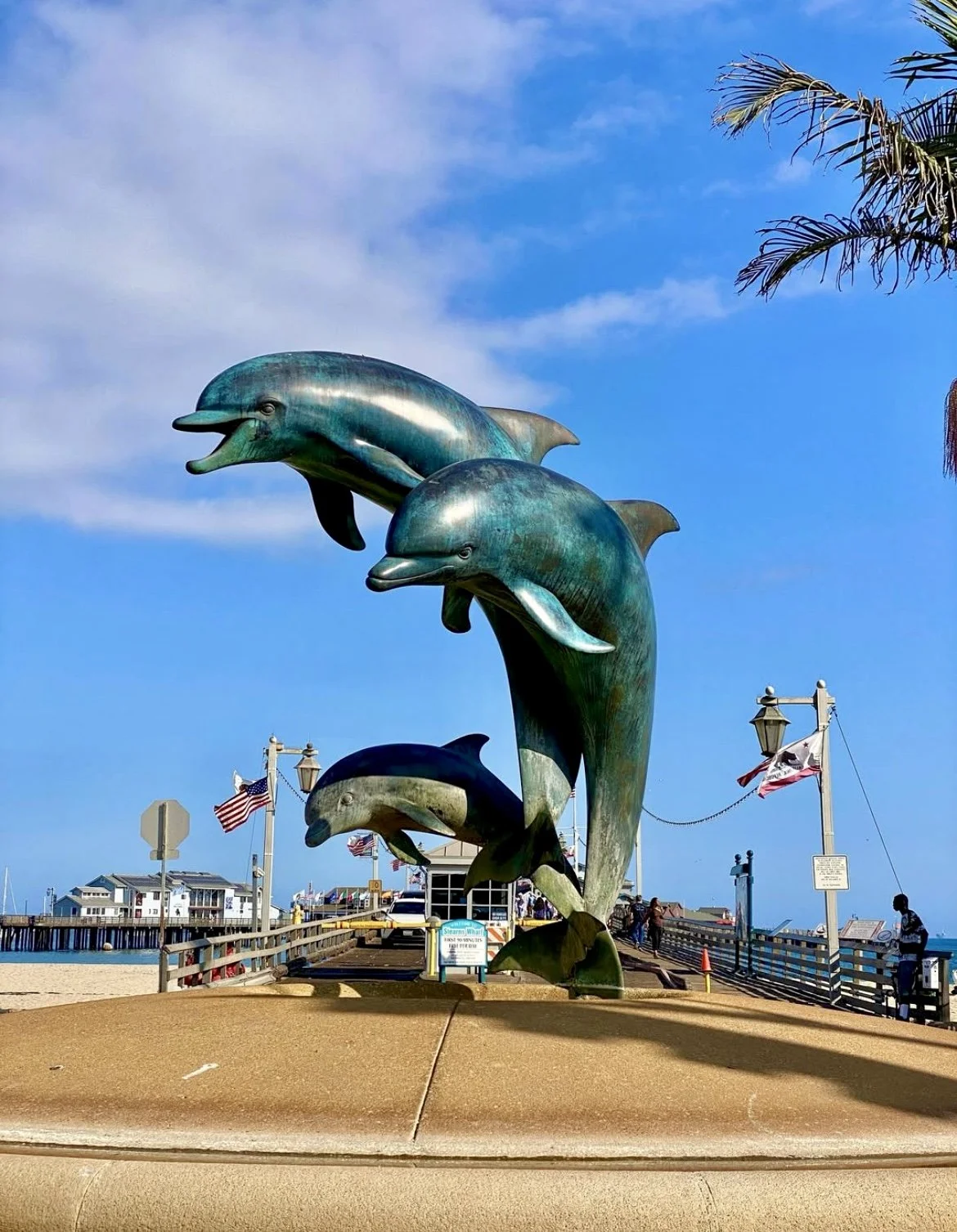 Bronze dolphin sculptures on a pier with people, flags, and buildings in the background under a blue sky.