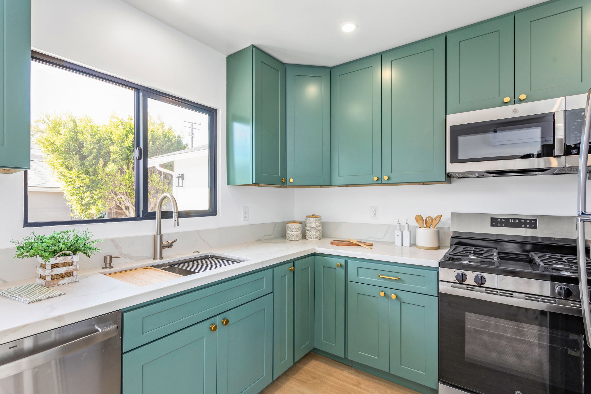 Kitchen with turquoise cabinets, white marble countertops, stainless steel appliances, and a large window letting in natural light.