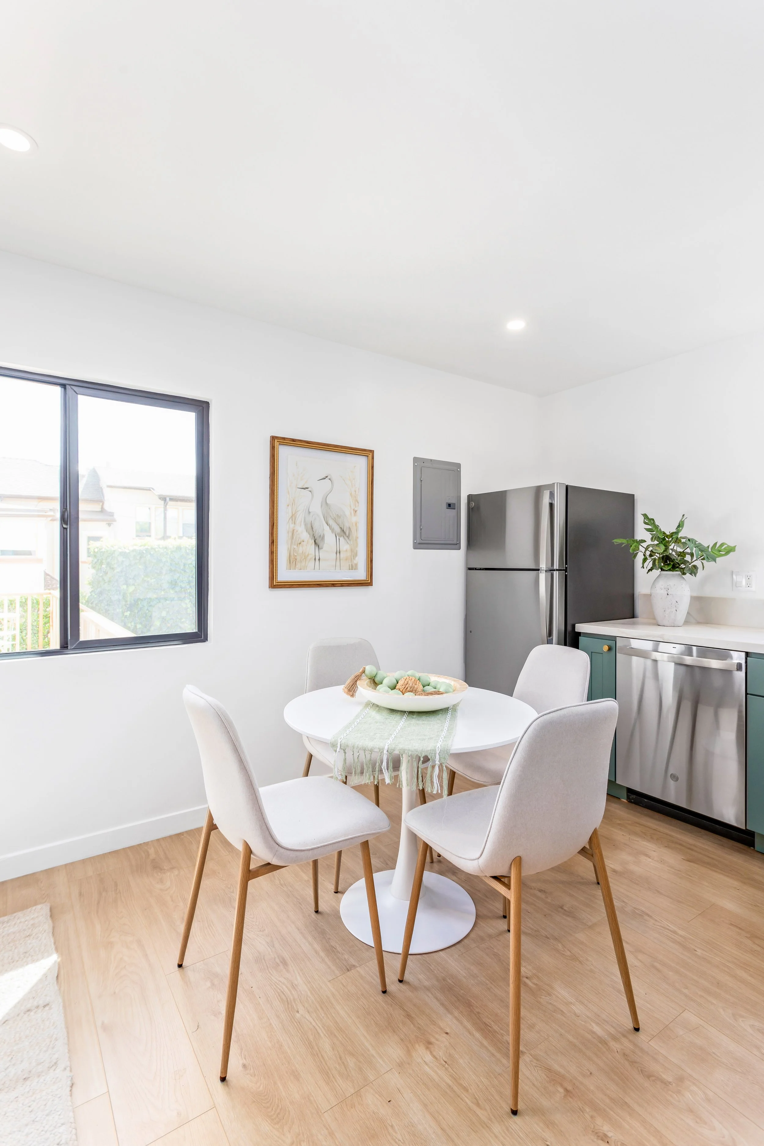 A bright dining area with a white round table, four beige chairs, a fruit bowl, and a window overlooking neighboring houses. Part of the kitchen with a refrigerator, a potted plant, and countertop is visible.