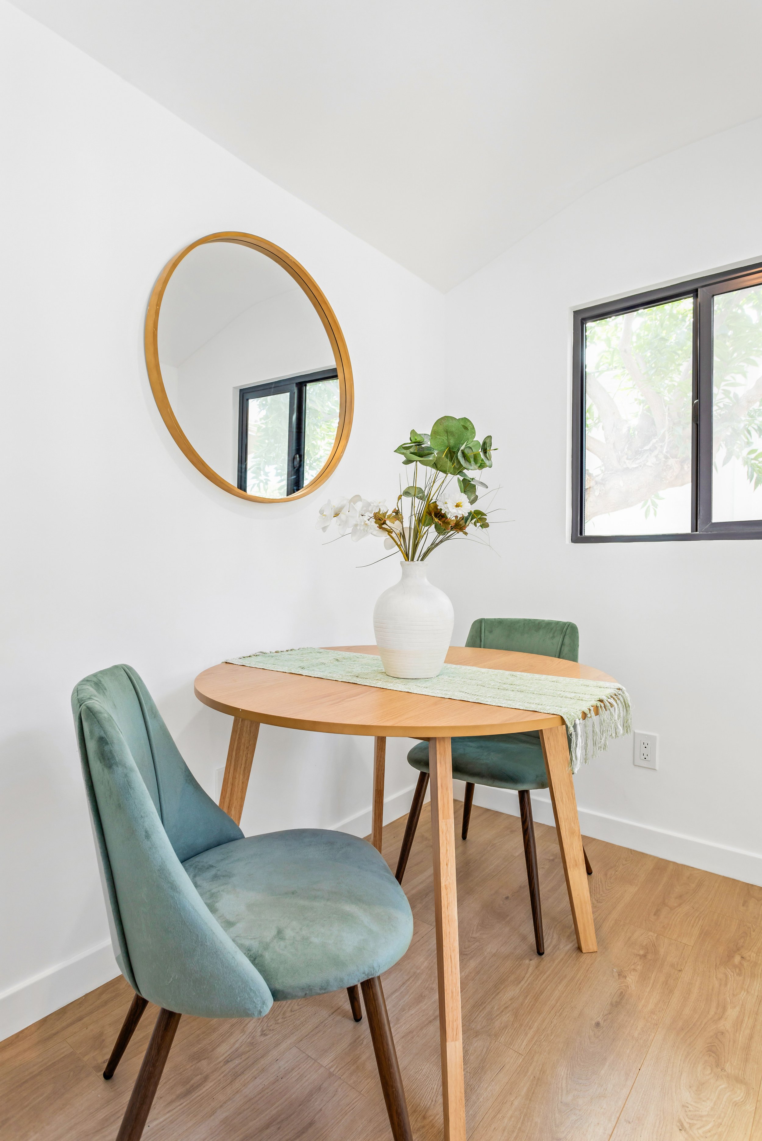 A dining area with a wooden table, two green upholstered chairs, a vase with green and white flowers, a round mirror on the wall, and a window showing greenery outside.