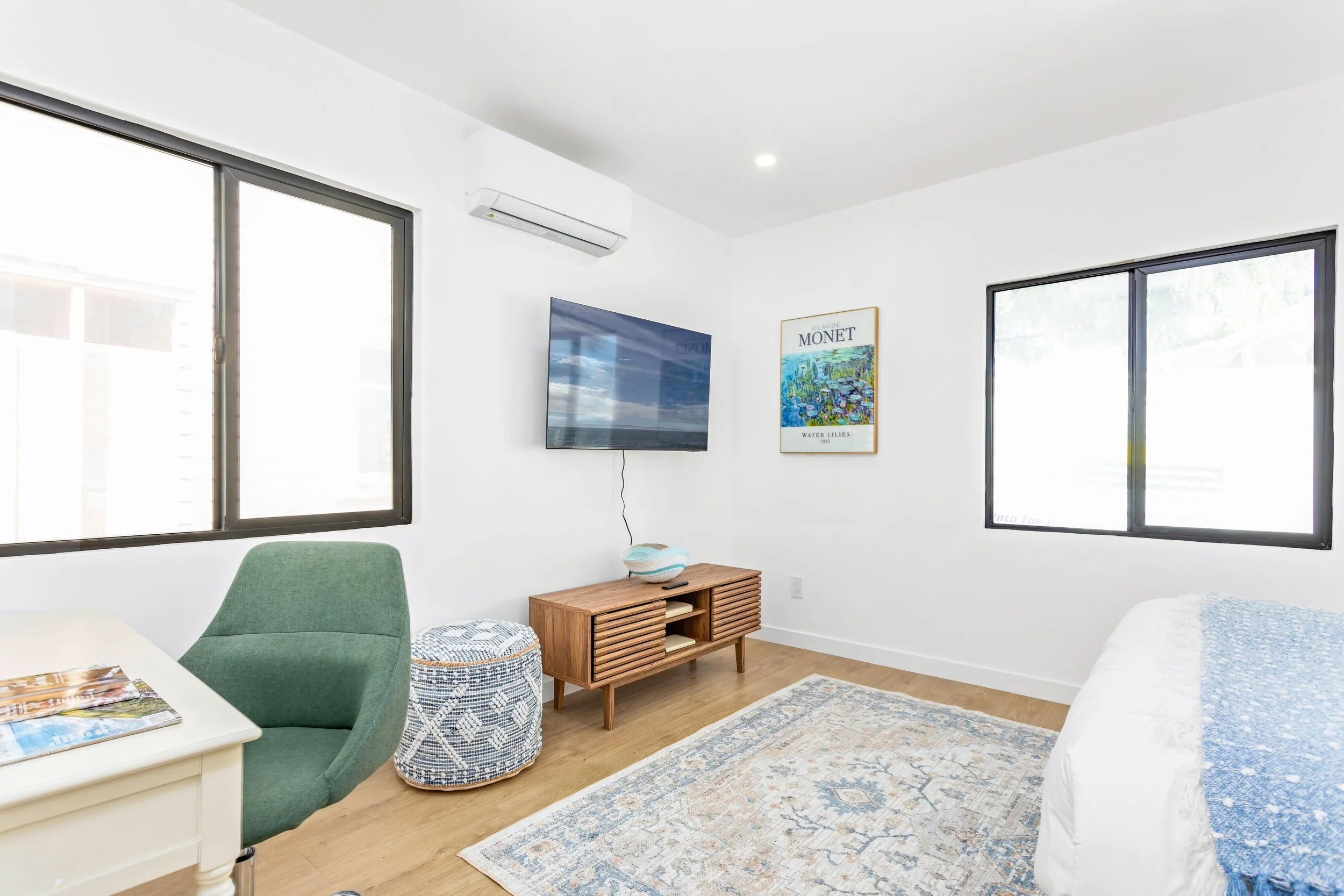 Bright living room with white walls, two large windows, a TV on the wall, a green armchair, a wooden media console, a round basket, a framed Monet art print, a white bed with a blue blanket, and a patterned area rug.