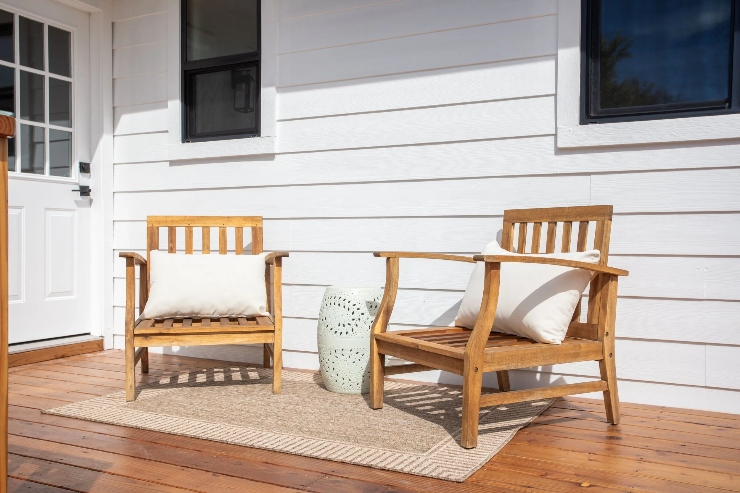 Outdoor patio with two wooden chairs with white cushions, a small white table, on a beige rug, with a white exterior wall and windows in the background.