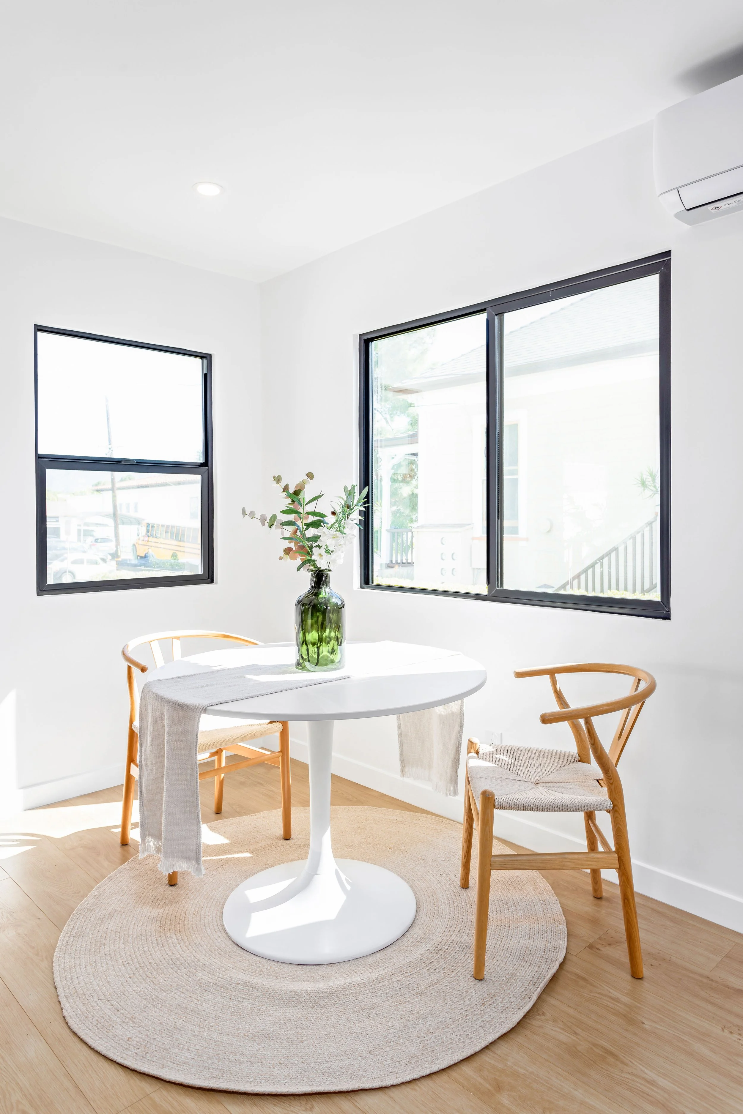 Sunlit dining area with a white round table, two wooden chairs, a green glass vase with flowers, and a beige rug on light hardwood floor, with large windows showing outside view.