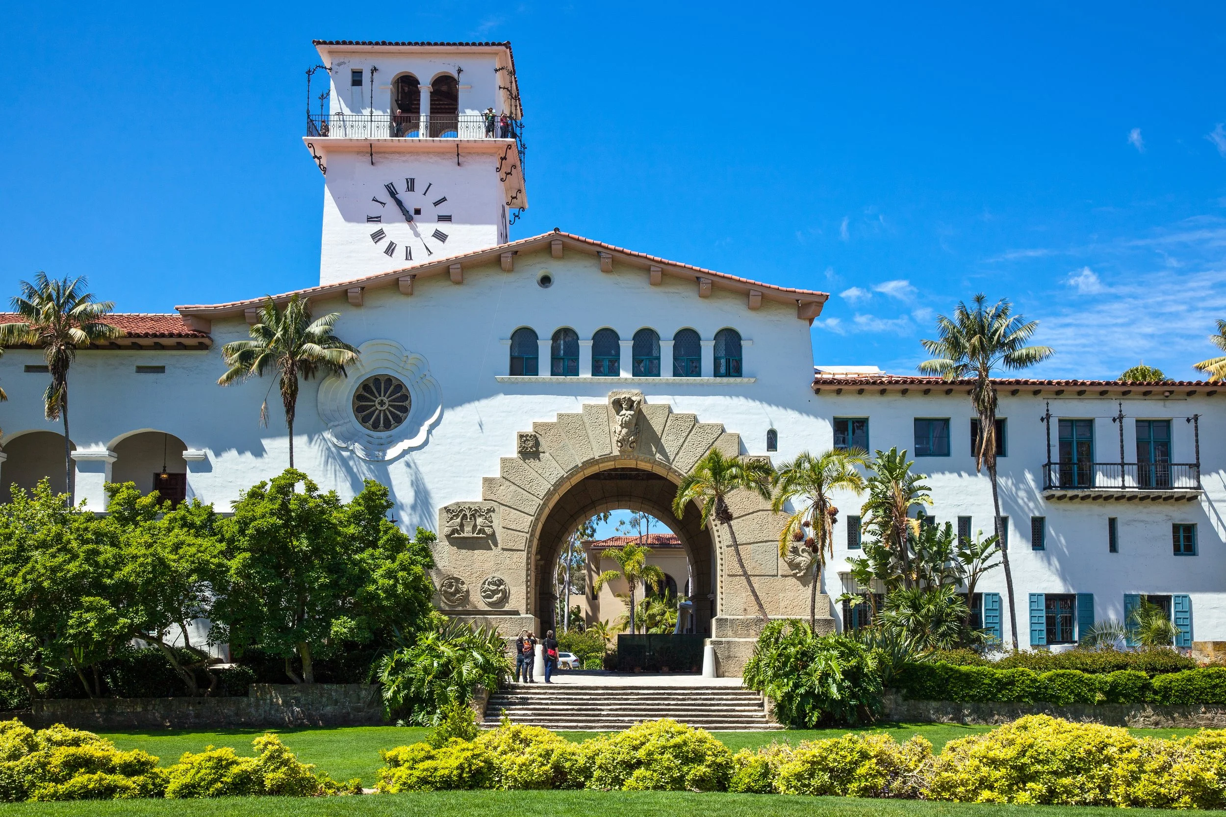 The image shows a historic white building with a clock tower and a large archway entrance, surrounded by palm trees and green bushes on a sunny day with a bright blue sky.