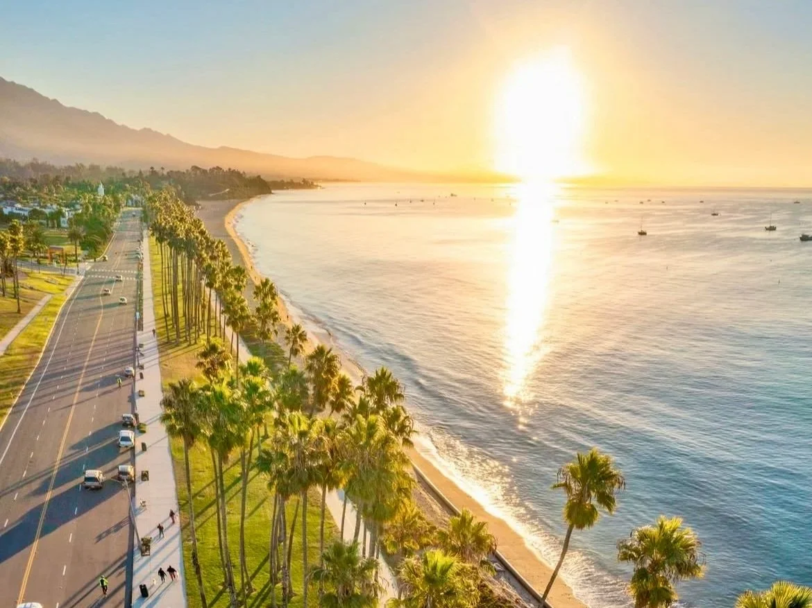 A coastal road runs parallel to a sandy beach lined with tall palm trees, overlooking a calm ocean with small boats, under a bright, setting sun with mountains in the background.