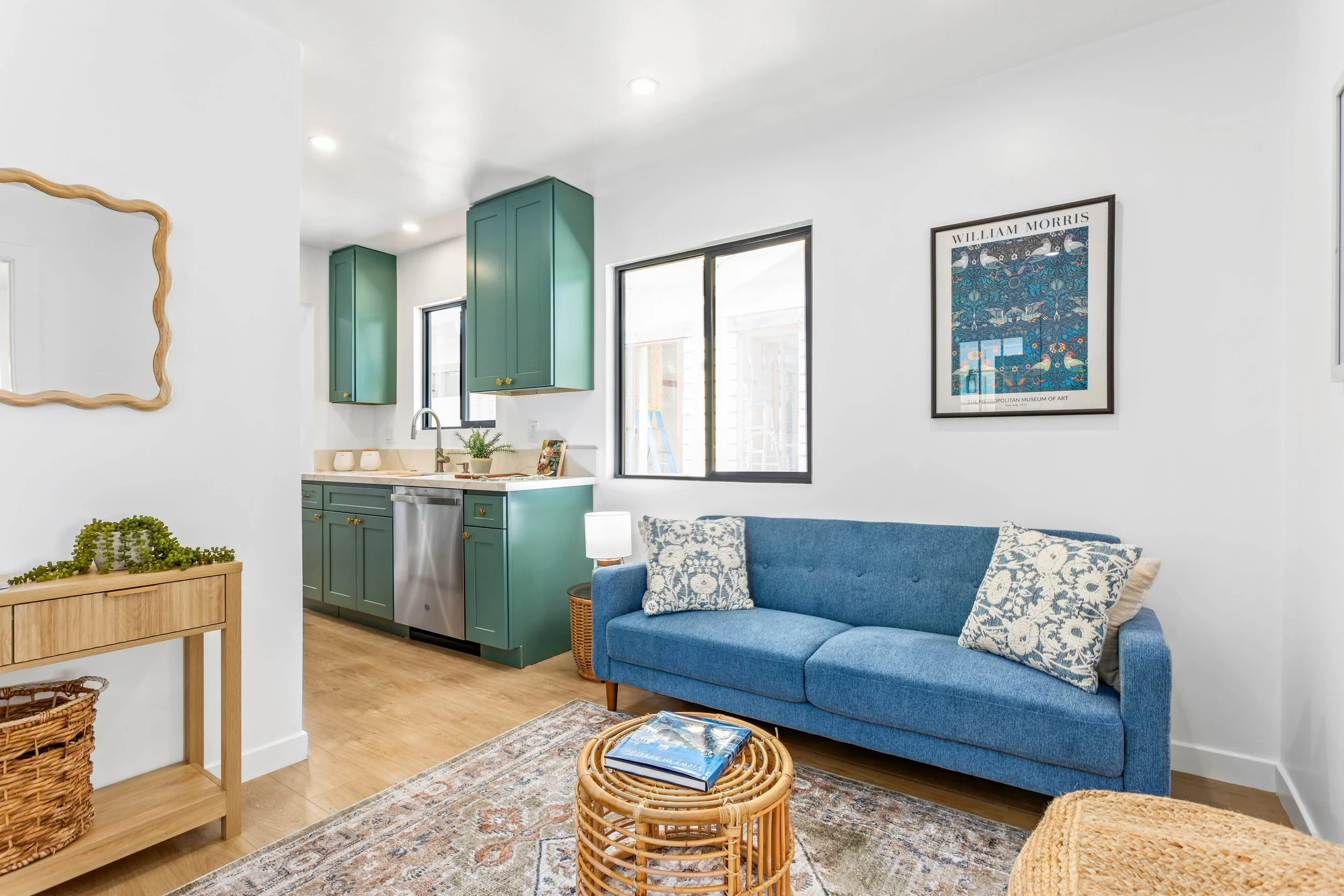 Living room with blue sofa, patterned pillows, wicker coffee table, and kitchen with green cabinets and window.