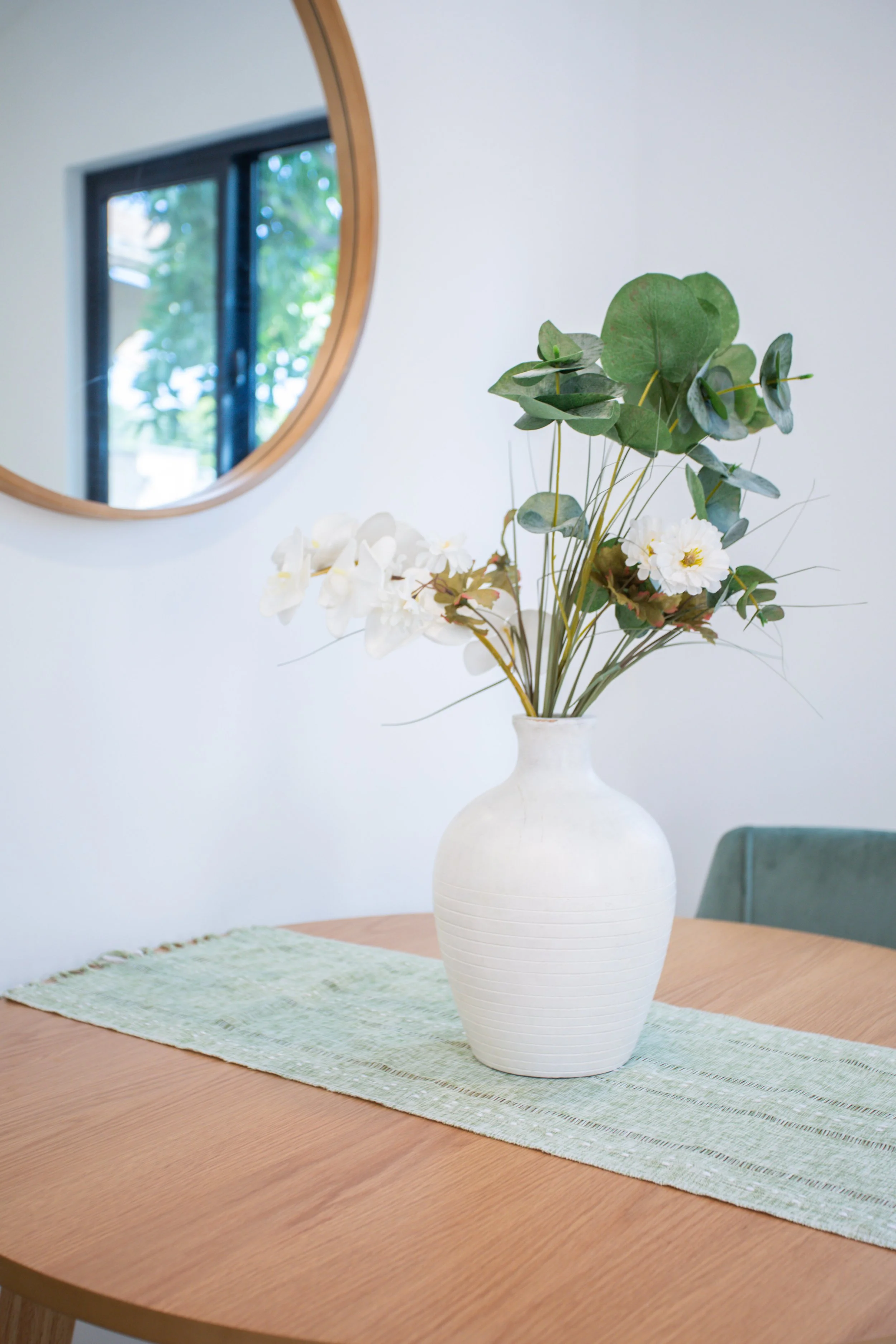 A white ceramic vase with green leaves and white flowers, set on a green table runner on a wooden dining table. A round mirror hangs on a white wall reflecting a window with trees outside.