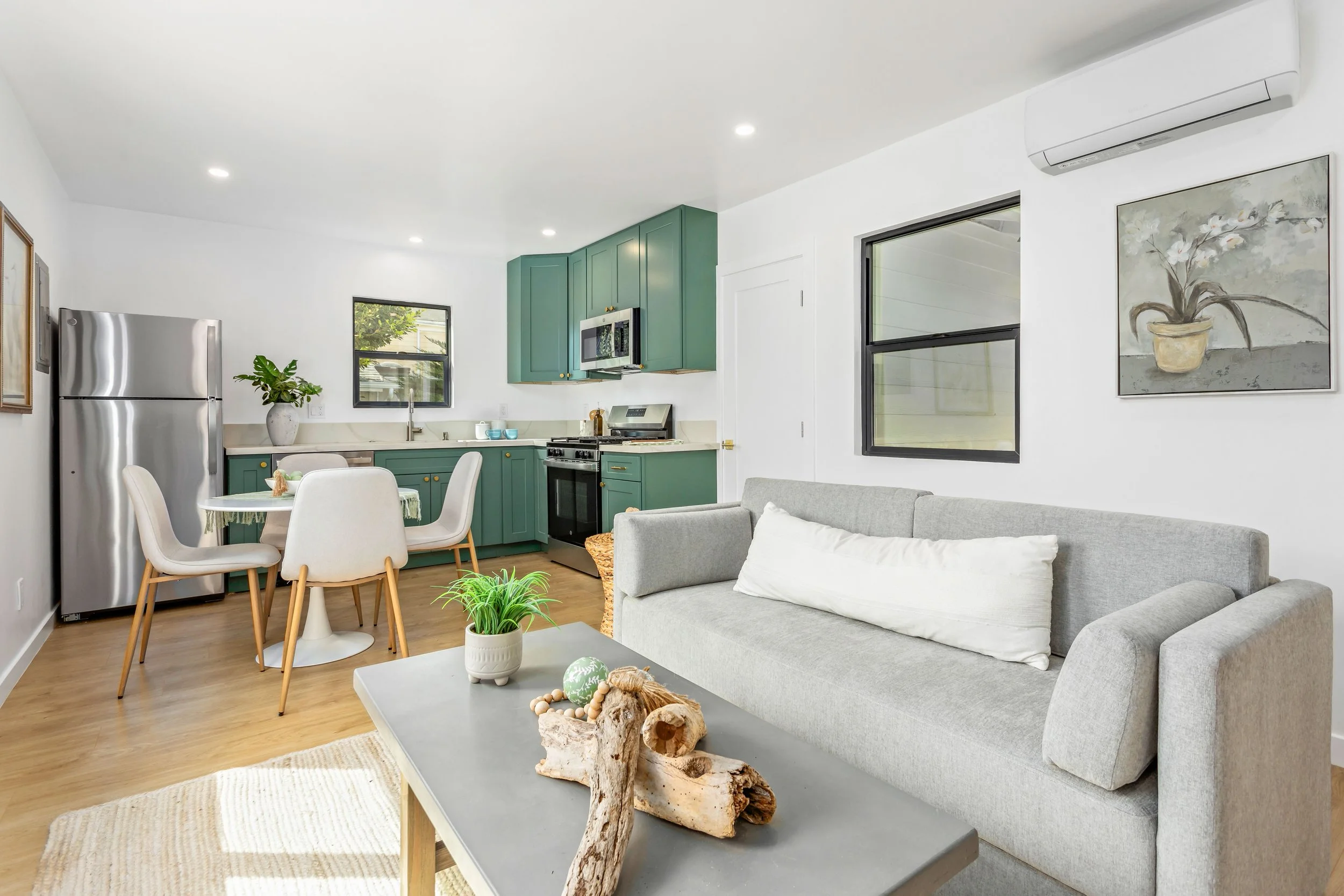 Open-concept living room and kitchen with gray sofa, green cabinets, dining table with white chairs, stainless steel refrigerator, and wall art, natural light through windows.