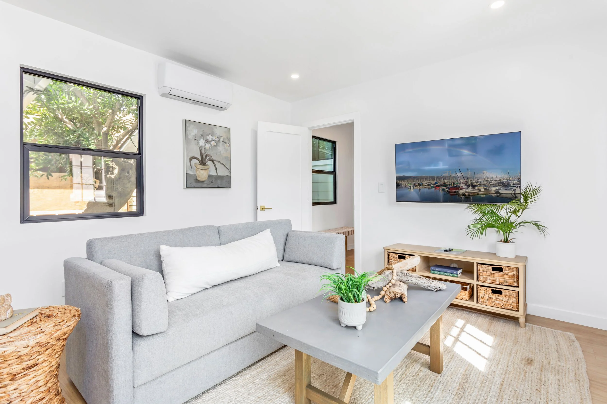 Living room with a gray couch, a light wood storage unit, a wall-mounted TV, a potted plant, windows showing greenery outside, and minimal decor.