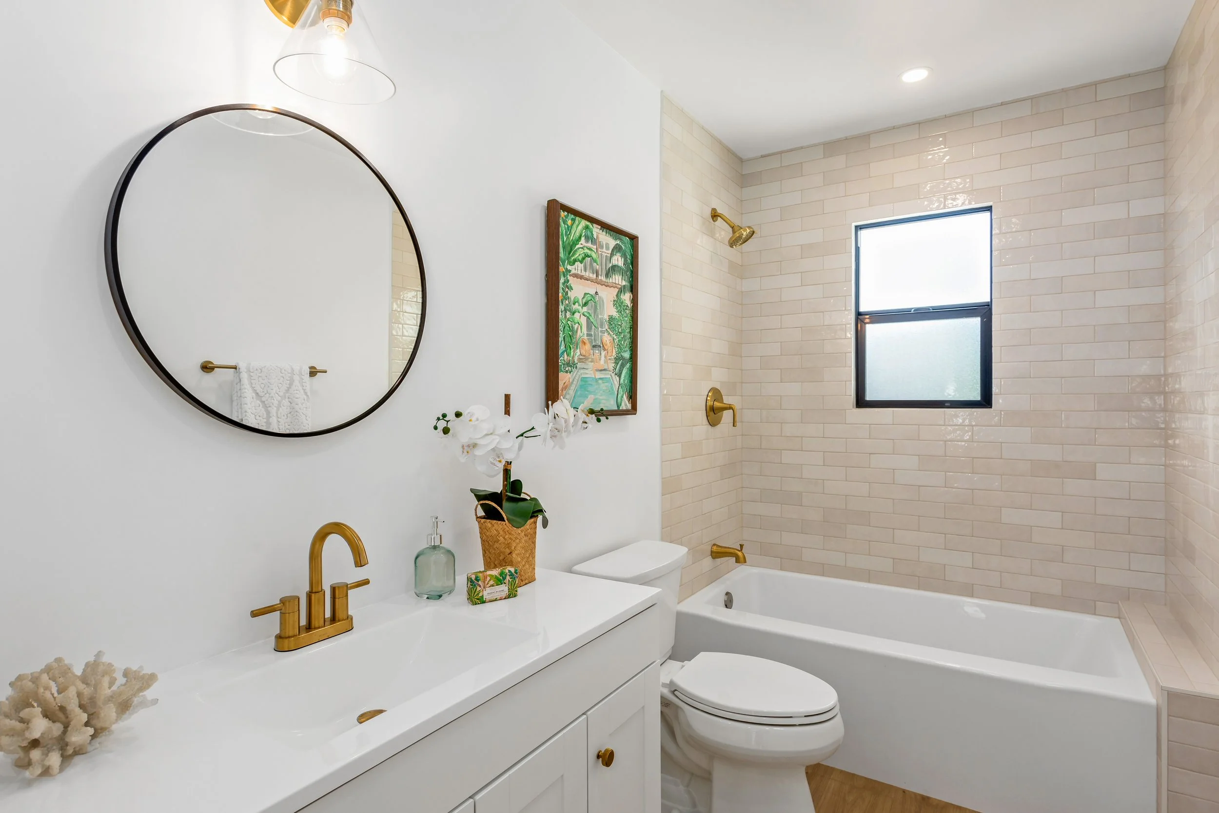 Modern bathroom with white vanity, round mirror, gold fixtures, white wall, tropical painting, orchid plant, coral decor, bathtub, beige subway tile walls, and frosted window.