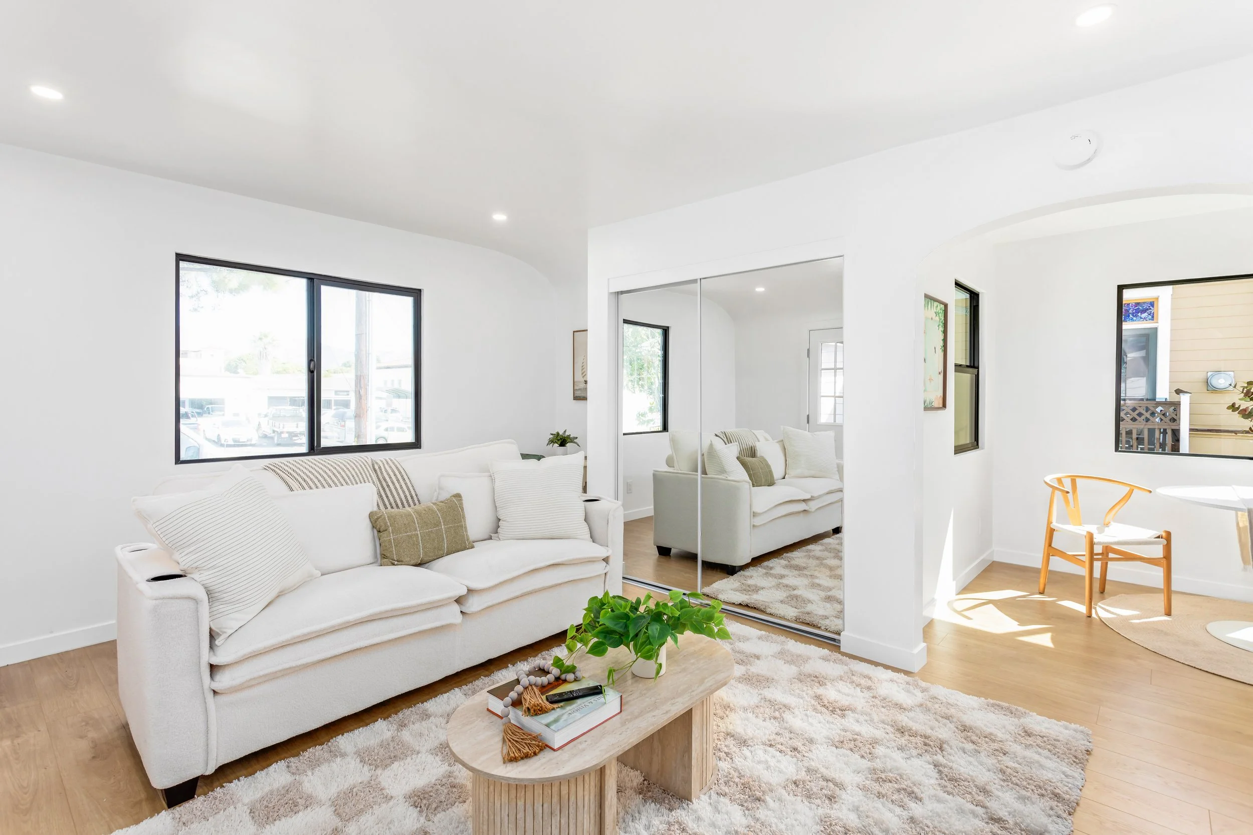 Bright living room with white walls, a white sofa with cushions, a wooden coffee table with a plant, books, and a tray, a large window, a mirrored closet, and a small dining area with a wooden chair and white table.
