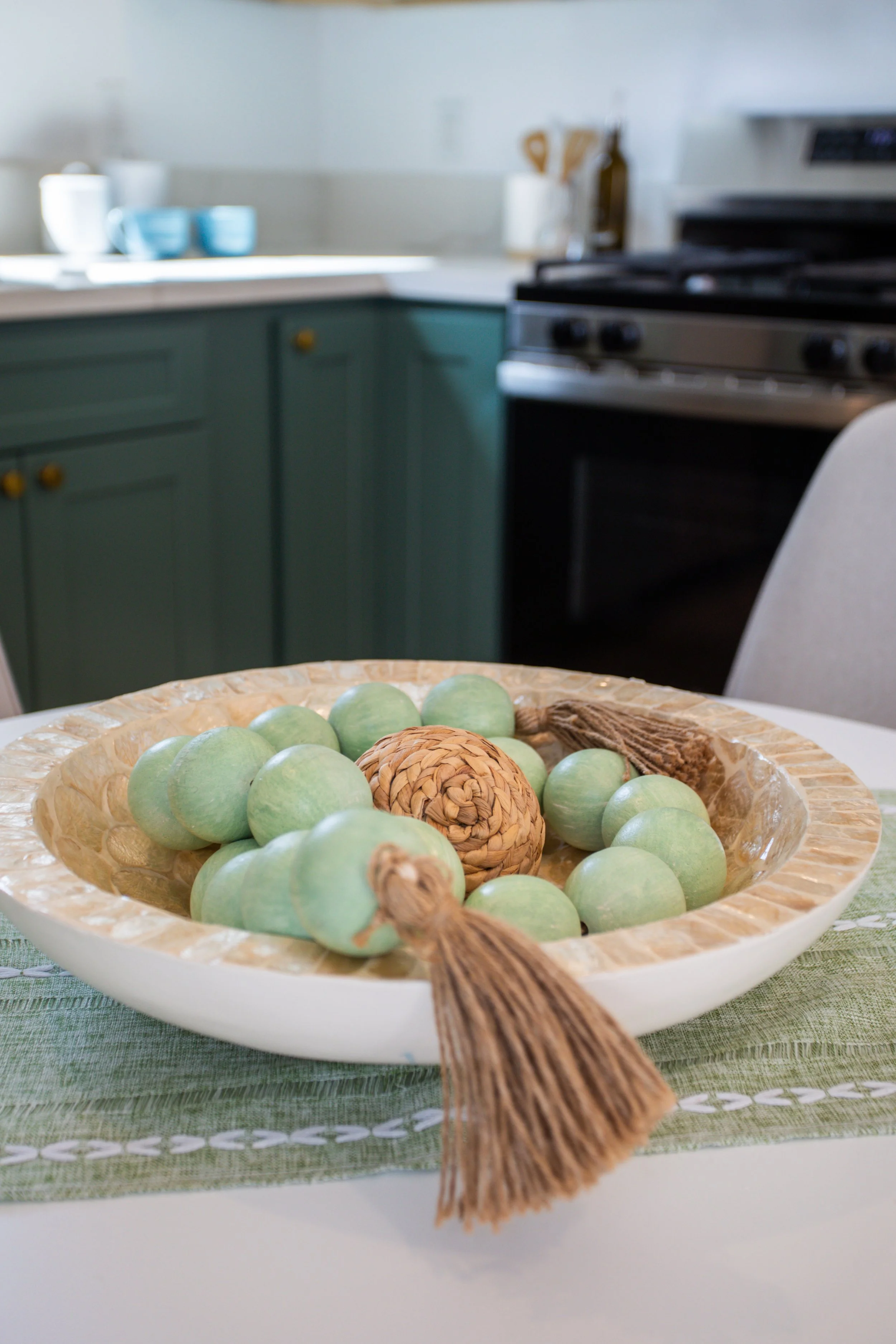 Decorative bowl filled with green wooden balls and a carved wooden sphere with tassels, placed on a dining table with a green placemat.