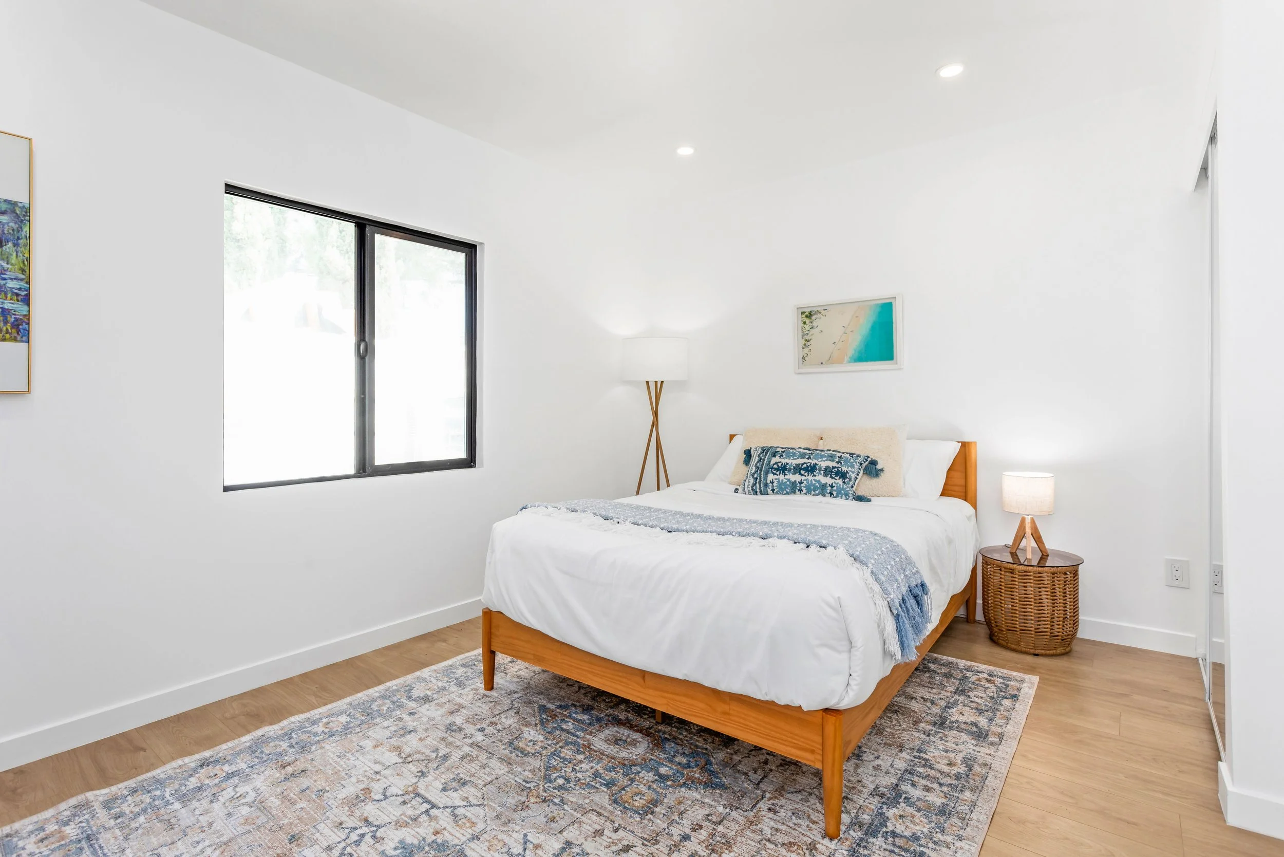 Bright bedroom with white walls, a wooden bed with white bedding, a blue decorative pillow, a wooden nightstand with a lamp, a floor lamp, a landscape painting, a window, and a patterned area rug.