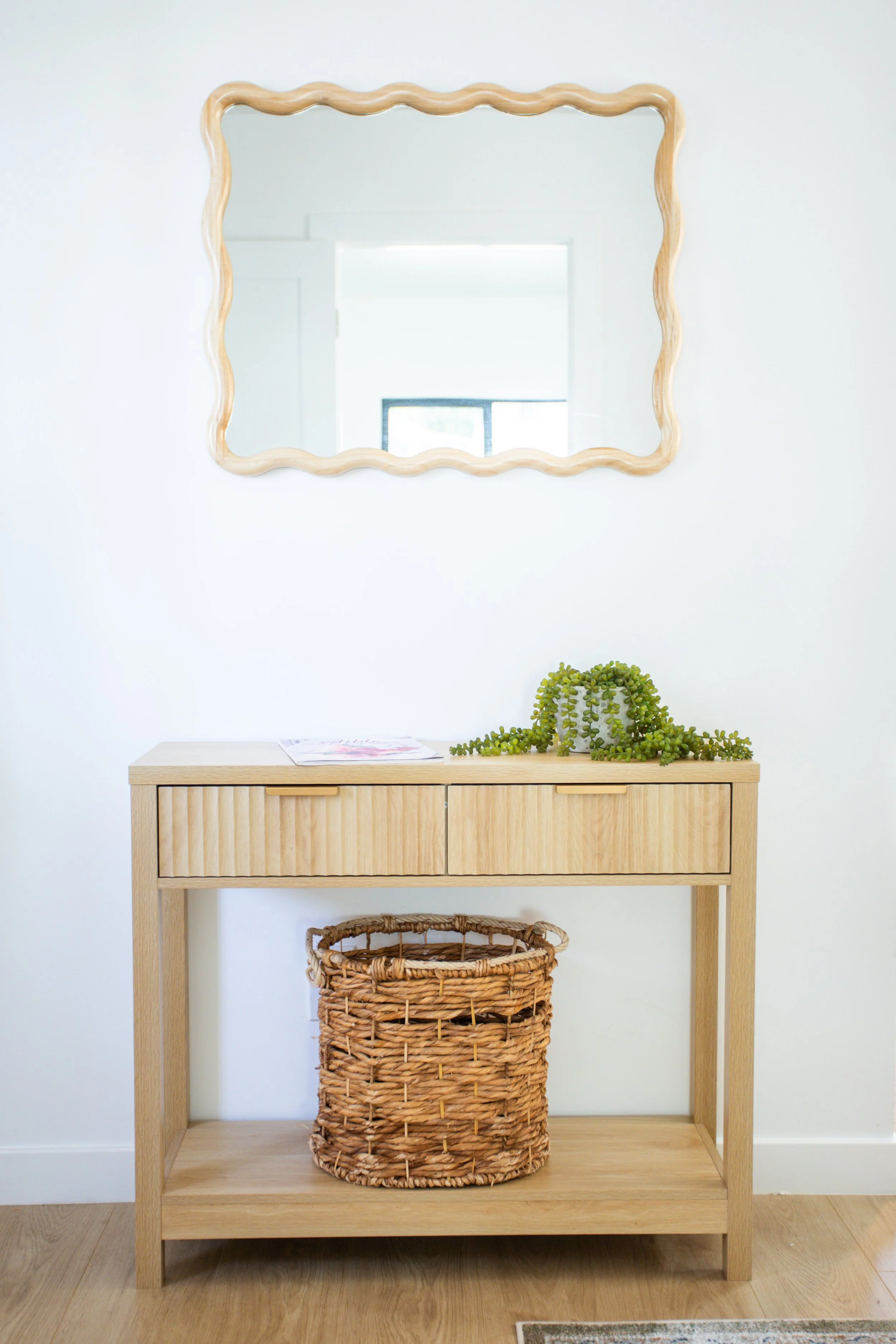 A wooden console table with two drawers and a lower shelf, a basket underneath, a green plant on top, a mirror with wavy wooden frame above, and a window reflected in the mirror, all set against a white wall.