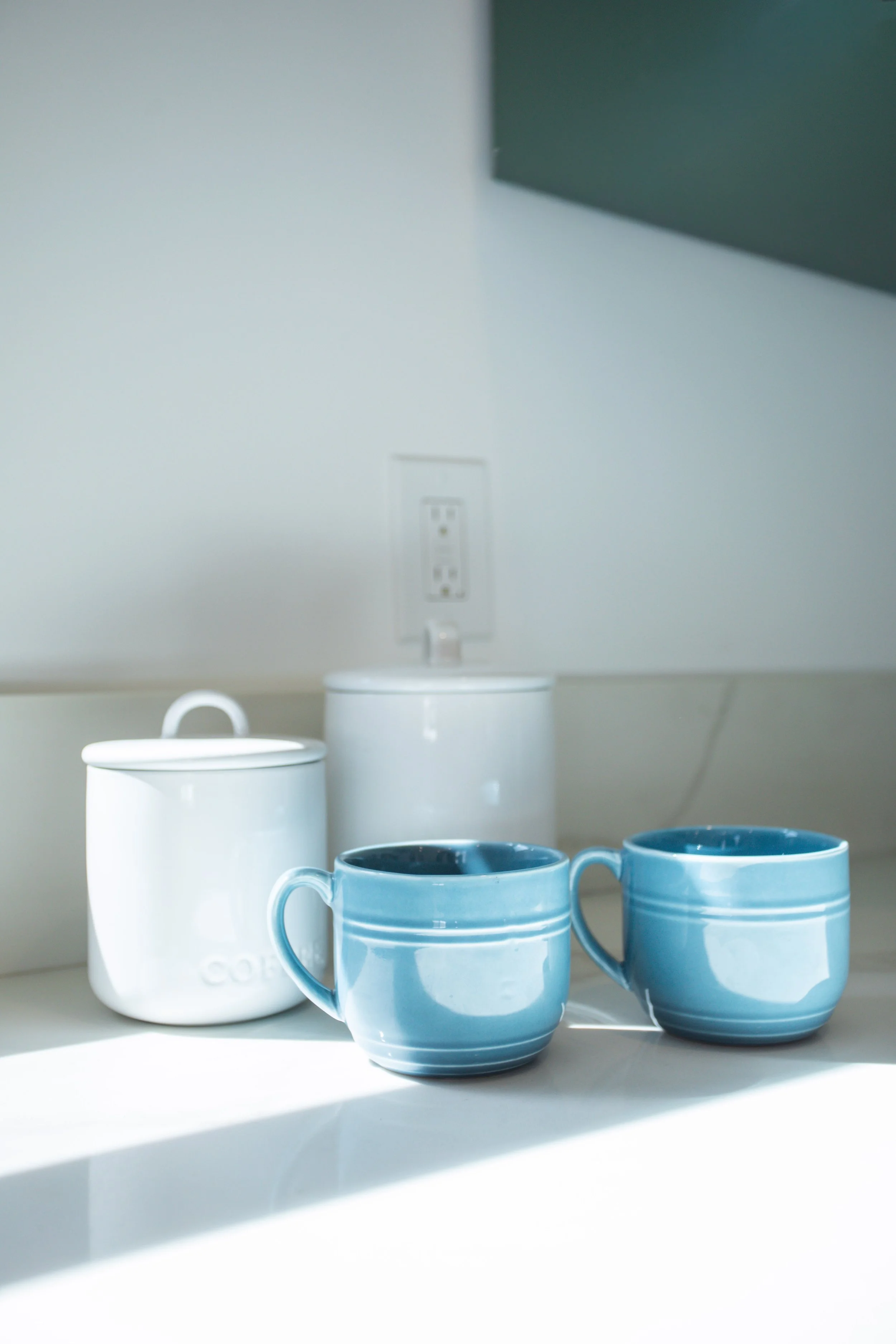Ceramic mugs and containers on a kitchen countertop, with a white wall and electrical outlet in the background.