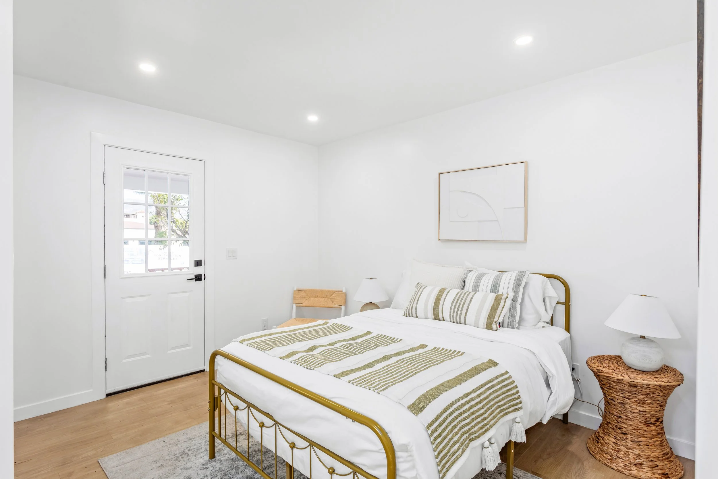 A bedroom with white walls, ceiling, and bedding, featuring a bed with a gold metal frame, decorative pillows, a wicker nightstand with a white lamp, and a windowed door showing an outdoor view.