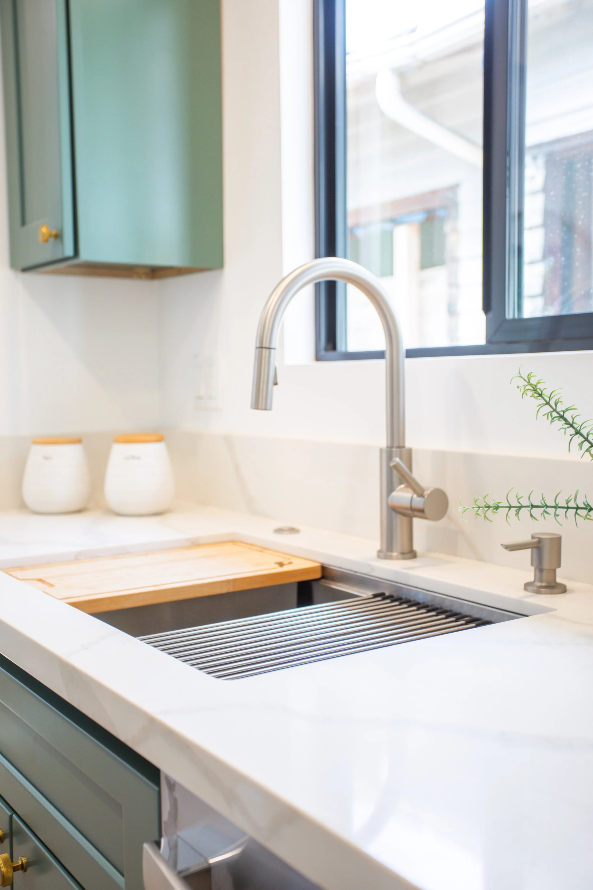 A modern kitchen sink with a stainless steel faucet and a drain cover, a window above, and green cabinets and decor.