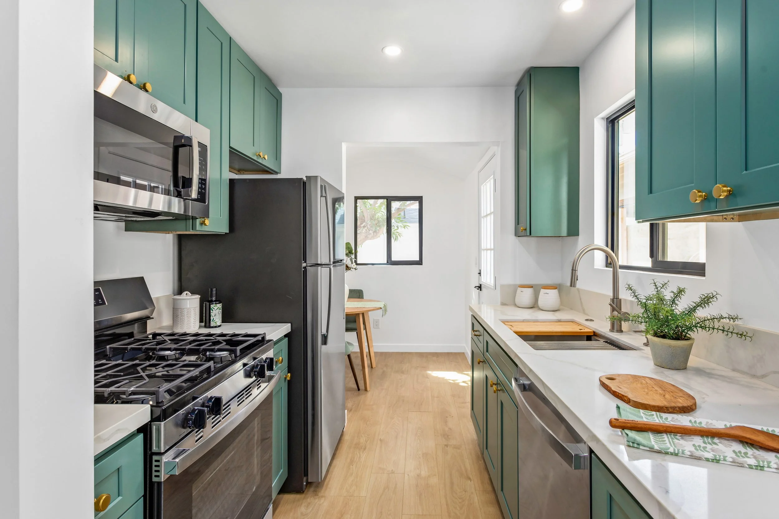 Modern kitchen with green cabinets, stainless steel appliances, wooden flooring, and a white counter with potted plant and cutting boards.