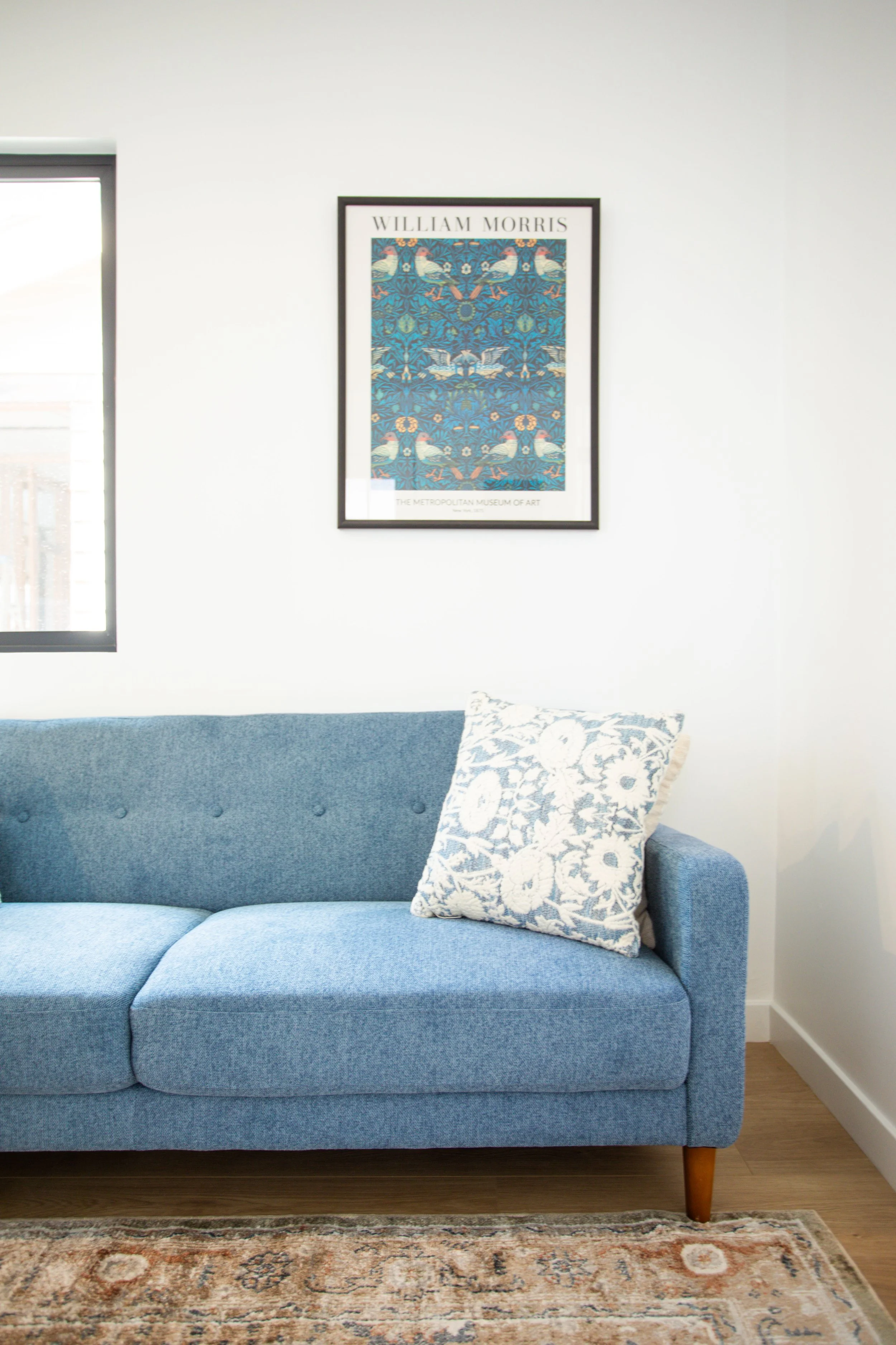 Interior of a living room with a blue sofa, floral cushion, wall art above the sofa, and a window to the left.