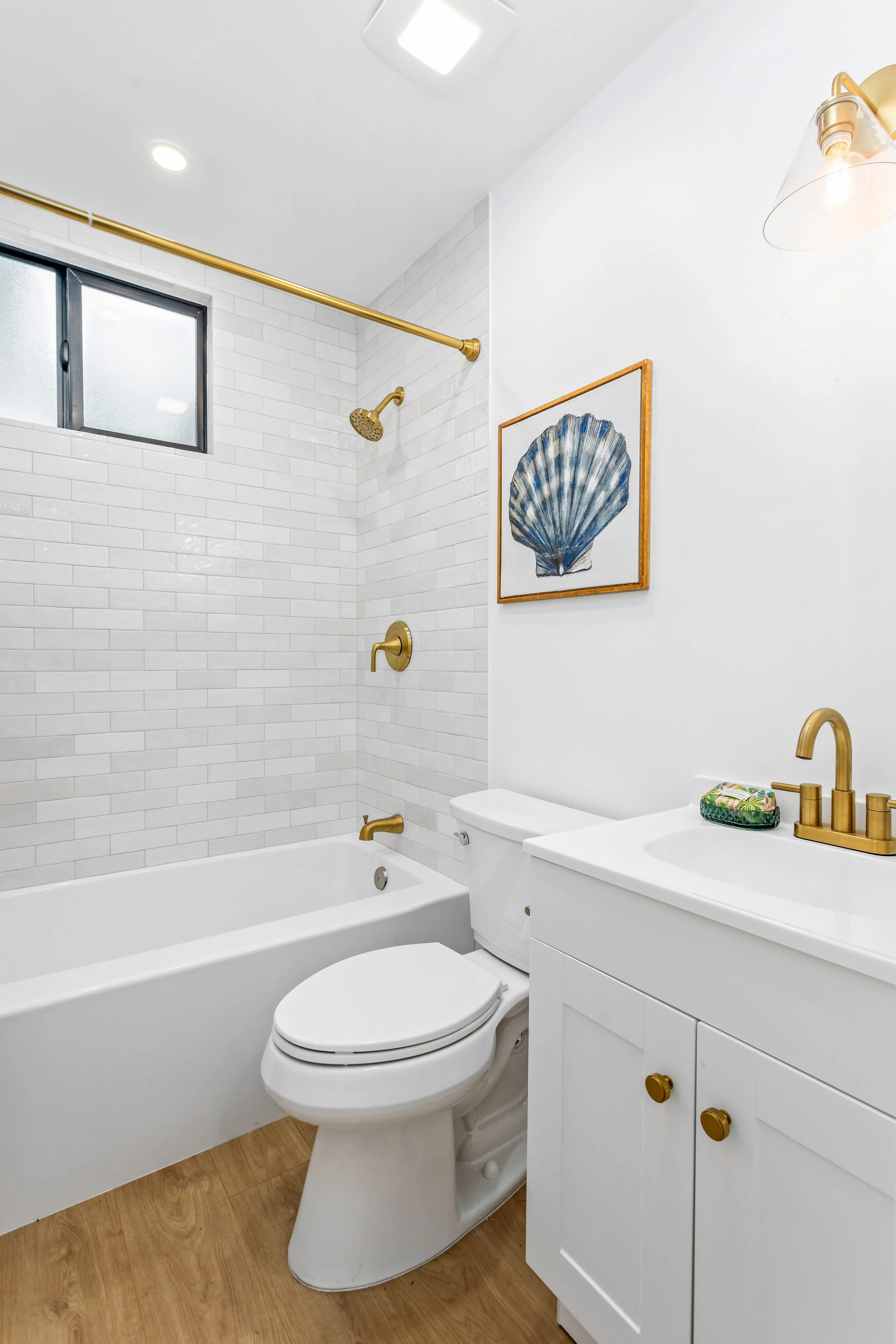 A modern bathroom with white subway tile walls, a bathtubs, a white toilet, a white vanity with gold fixtures, a framed picture of a seashell on the wall, a small window, and wood flooring.