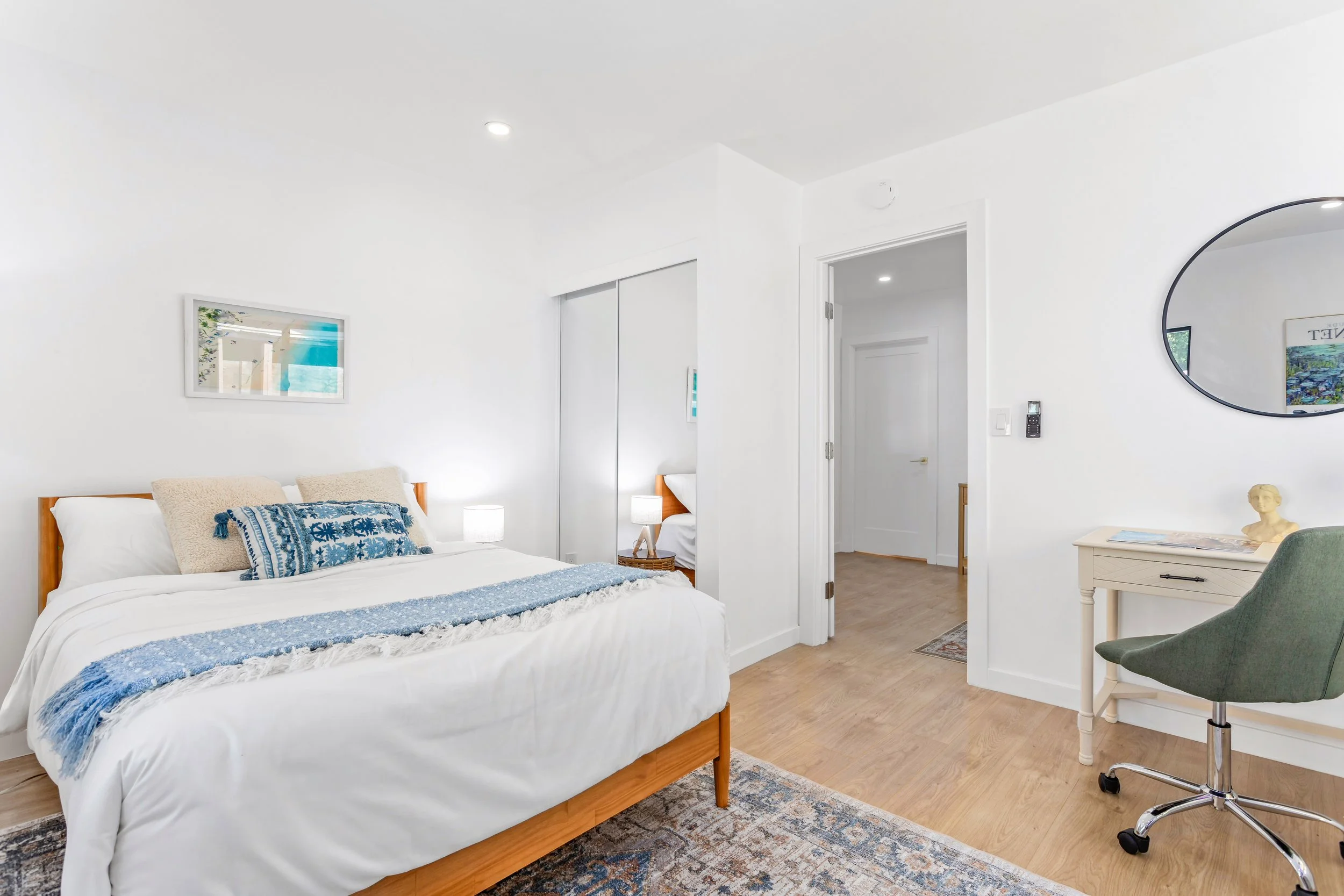 Clean bedroom with white walls, wooden bed with white bedding and blue accent pillows, small tables with lamps on each side, a mirror on the wall, a green desk chair, and a closet with mirrored doors.