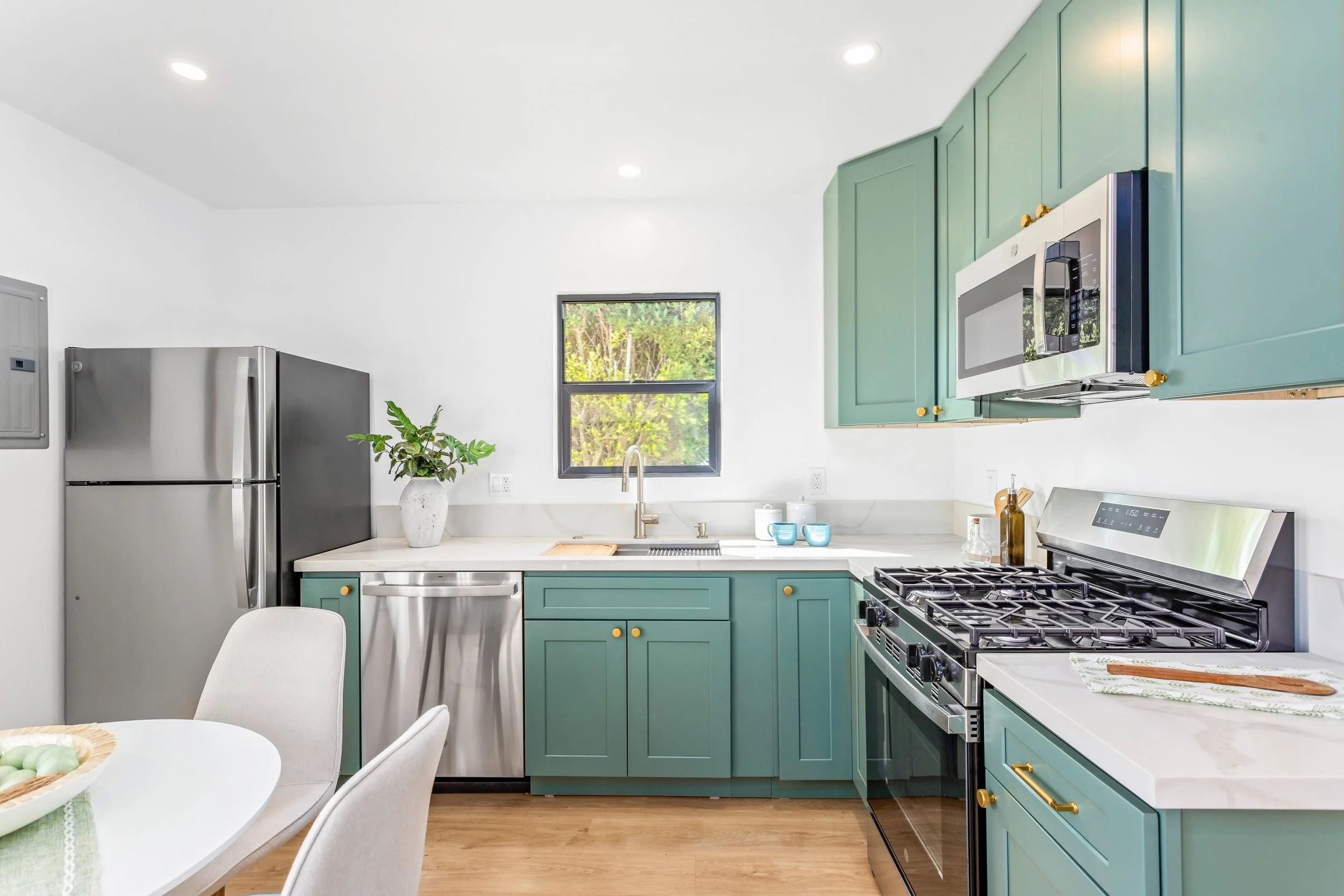 Modern kitchen with teal cabinets, stainless steel appliances including a refrigerator and a stove, a white countertop, a window above the sink, and a white dining table with chairs.