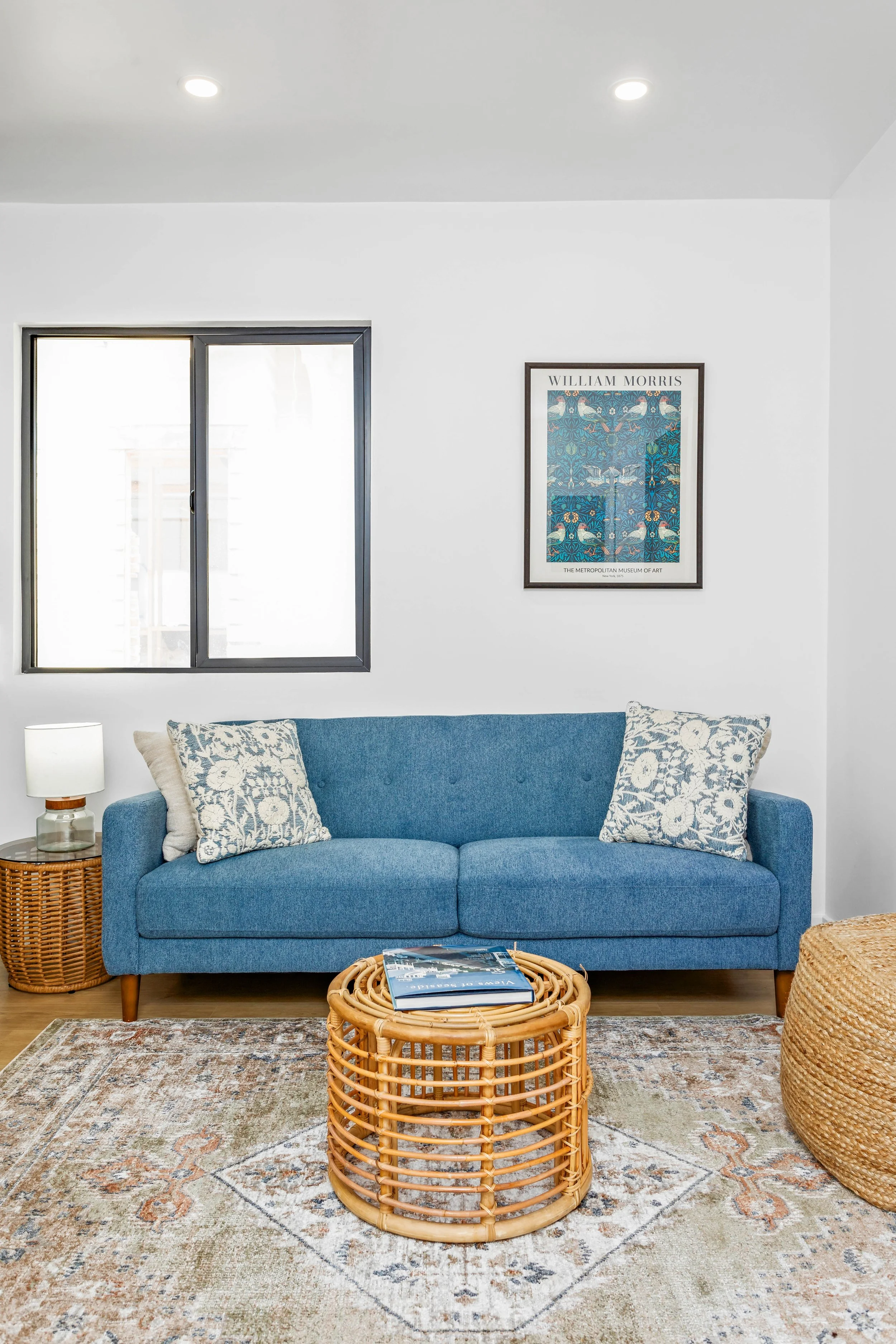 A living room with a blue sofa, two floral pillows, a wicker side table with a lamp, a round wicker coffee table with magazines, a Chinese style rug, a window with black trim, and a framed art print on the wall.