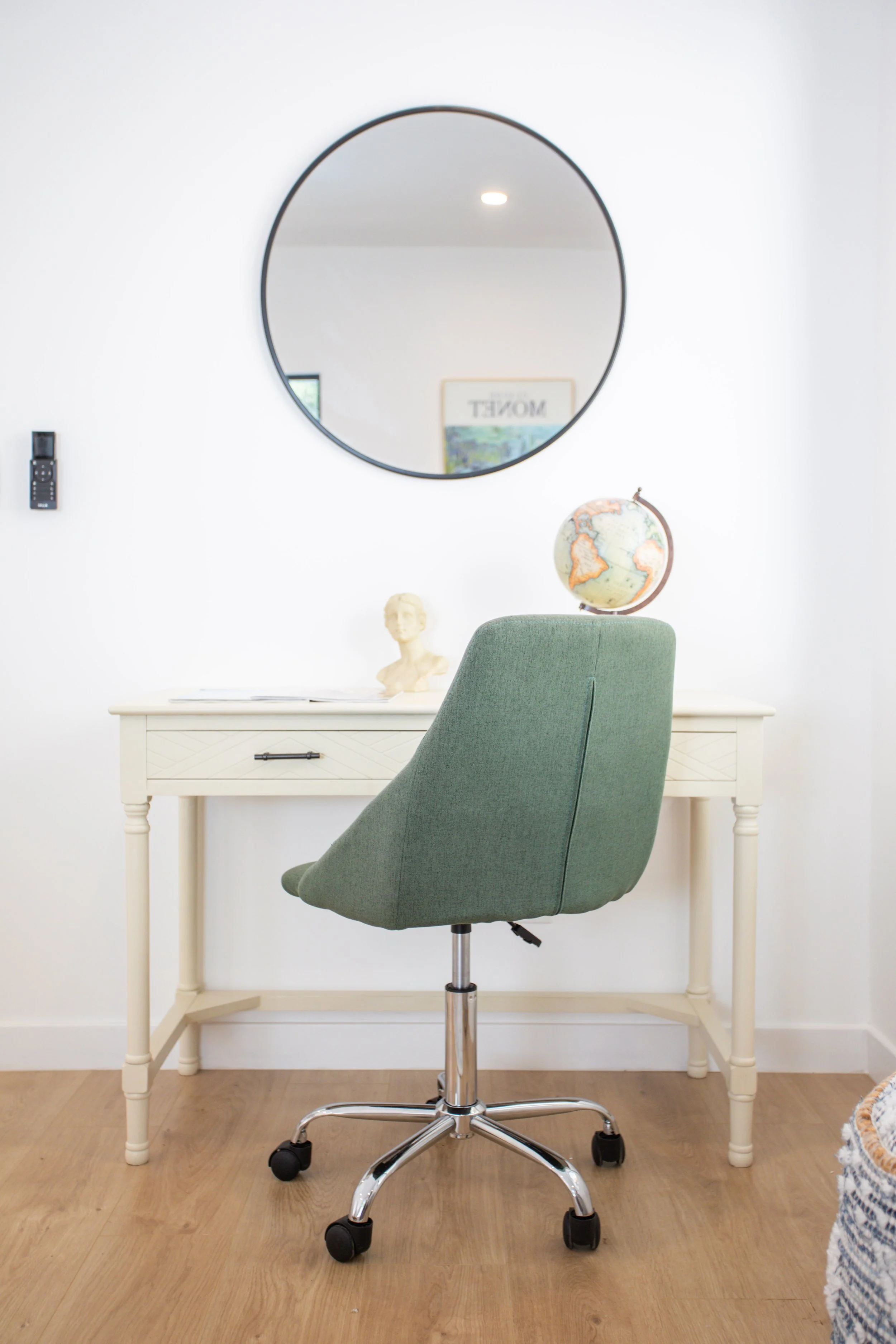 A white desk with a drawer, a bust sculpture, and a globe on top. A green office chair is in front of the desk, and a round mirror hangs on the wall above. The room has light-colored hardwood floors.
