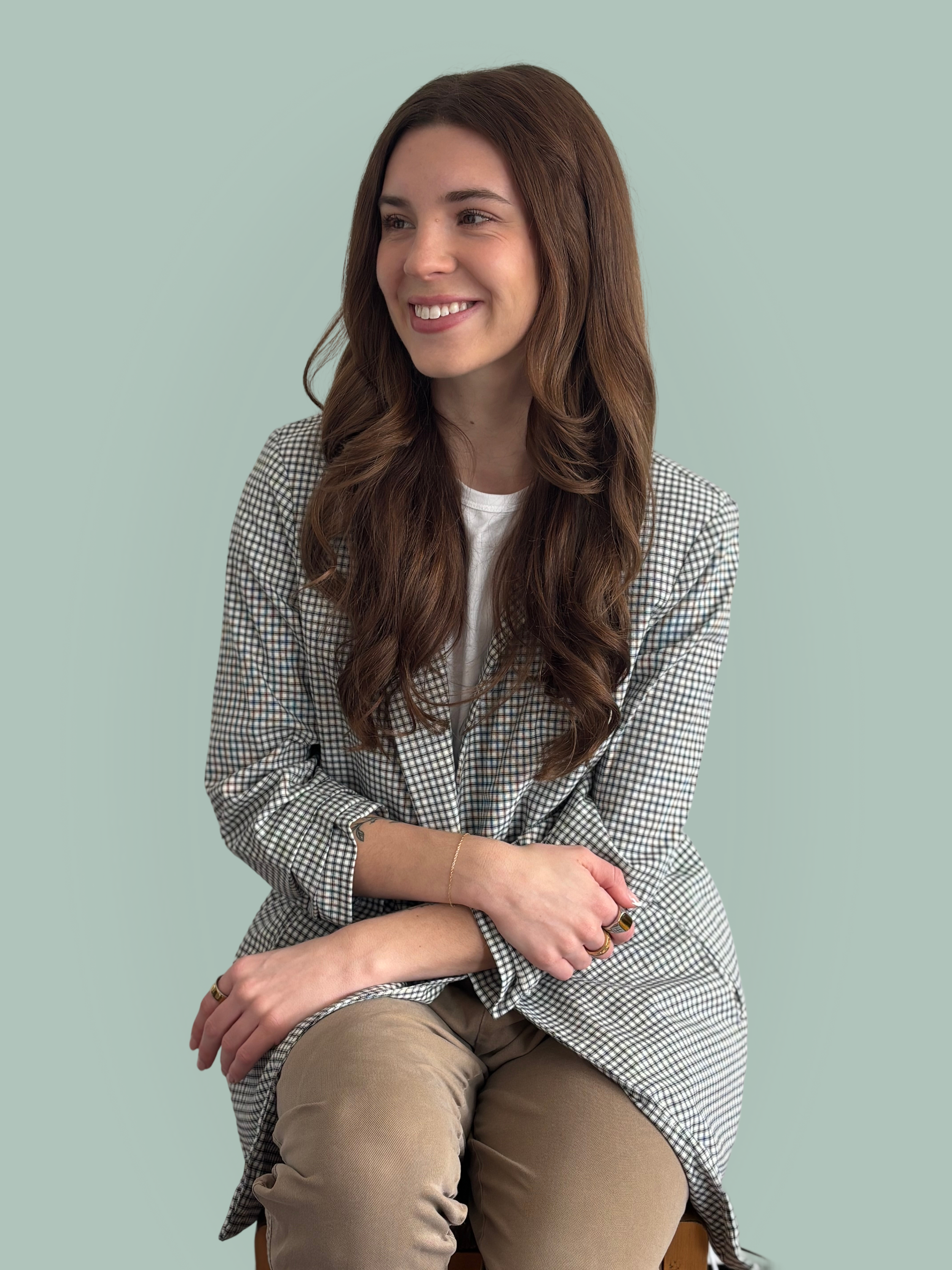 A young woman with long, wavy brown hair, smiling, sitting on a stool against a light teal background. She is wearing a checked blazer over a white top and beige pants.