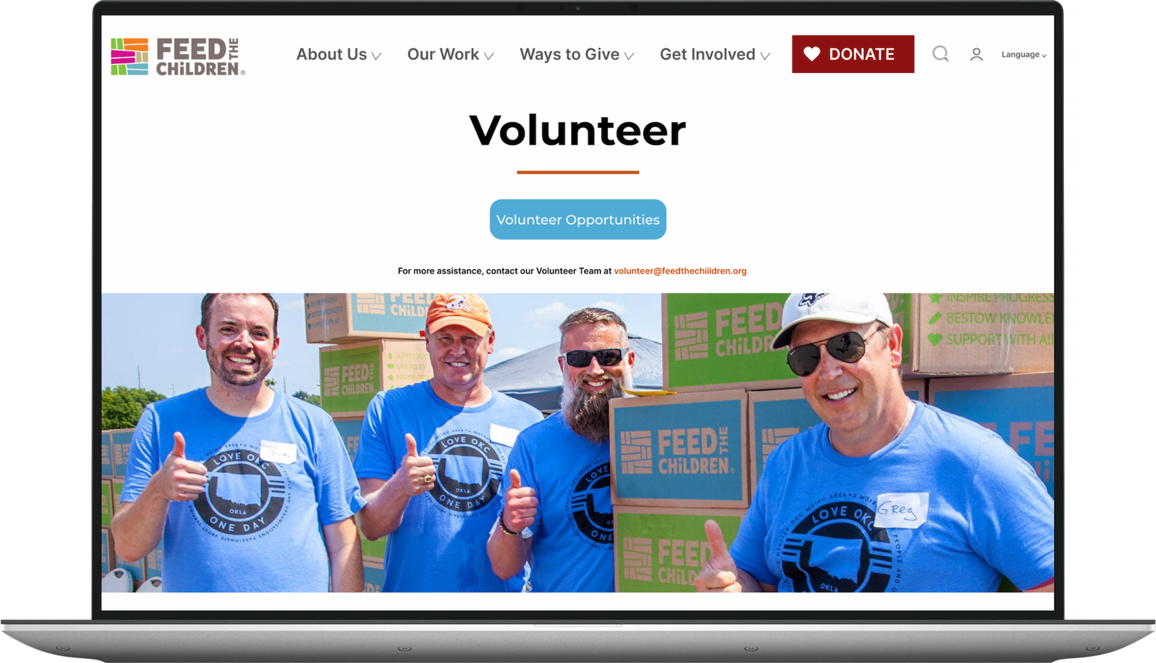 A group of four smiling volunteers wearing blue t-shirts with the 'Love OKC' logo, participating in a charity event for Feed the Children. They are standing outdoors in front of stacked boxes labeled 'Feed the Children,' with some volunteers giving thumbs-up gestures.