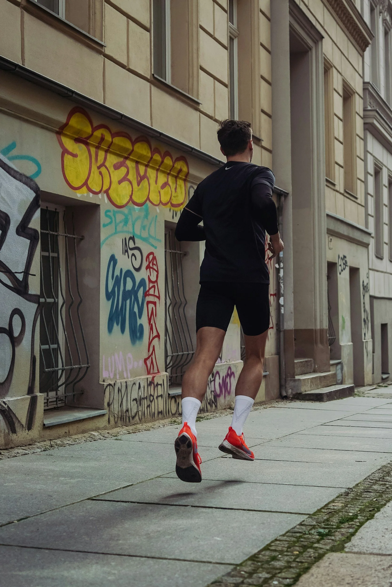Un homme court dans une rue urbaine, portant un t-shirt noir, des shorts noirs, des chaussures de course rouges et des chaussettes blanches, devant un mur graffiti.