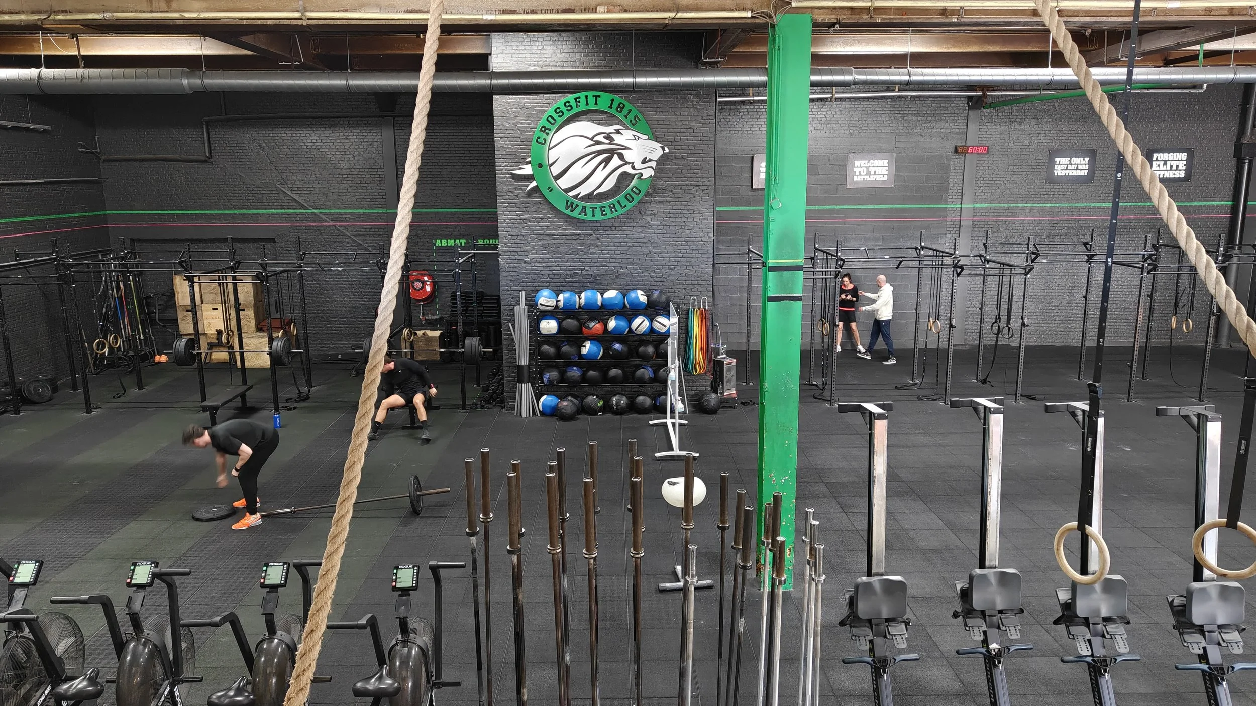 Une salle de sport avec plusieurs personnes s'entraînant, équipement de musculation, sacs de sport, et un mur avec le logo 'CrossFit 1815 Waterloo'.