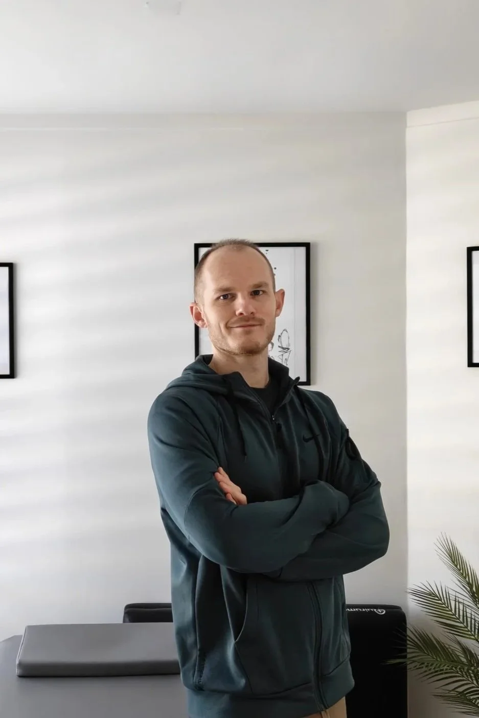 Un homme debout avec les bras croisés dans une pièce lumineuse, décoration avec des tableaux noirs et un plante verte.