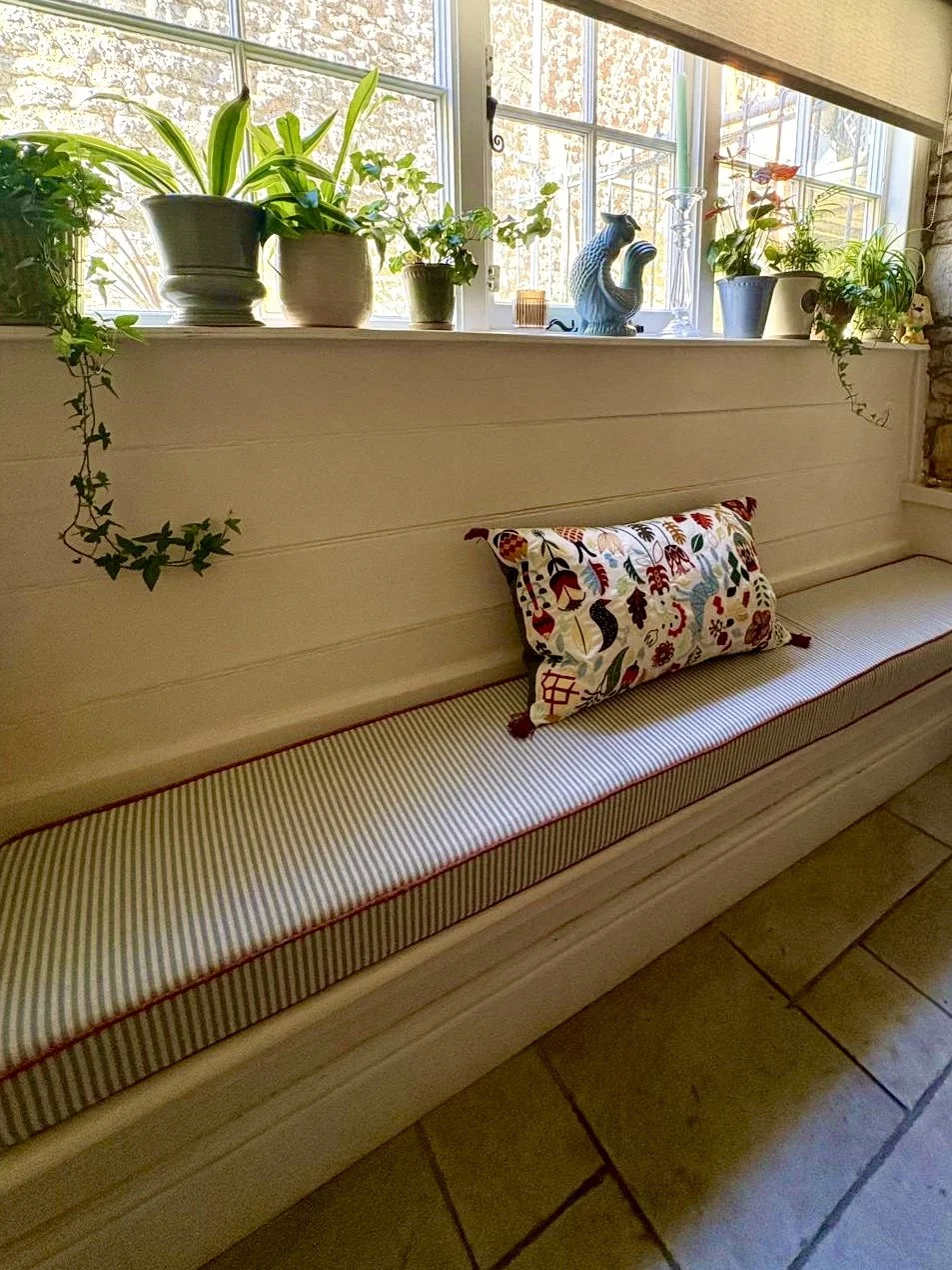 A window seat with a floral pillow and striped cushion, decorated with potted plants and figurines, with sunlight coming through the window.