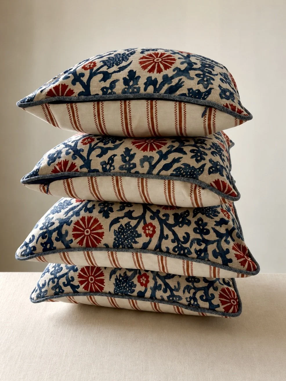 Stack of five decorative pillows with floral and striped patterns in red, blue, and white on a beige surface.