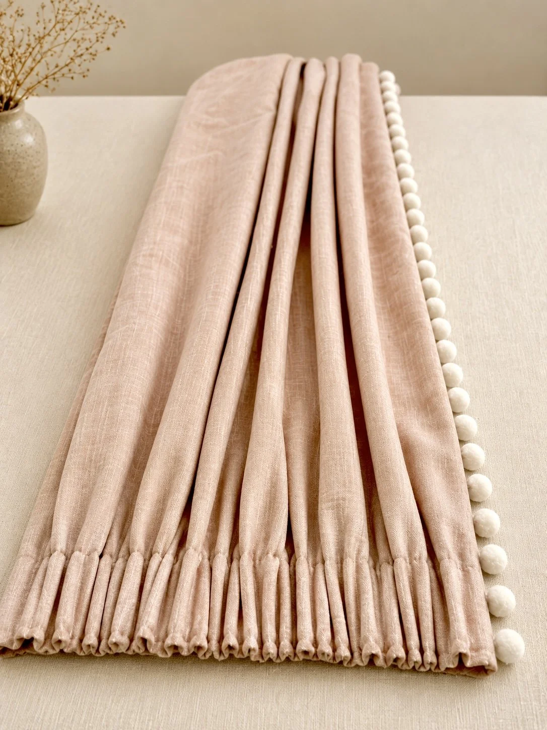 A soft pink curtain with white pom-pom trim, draped on a table with a beige vase containing dried flowers visible in the background.
