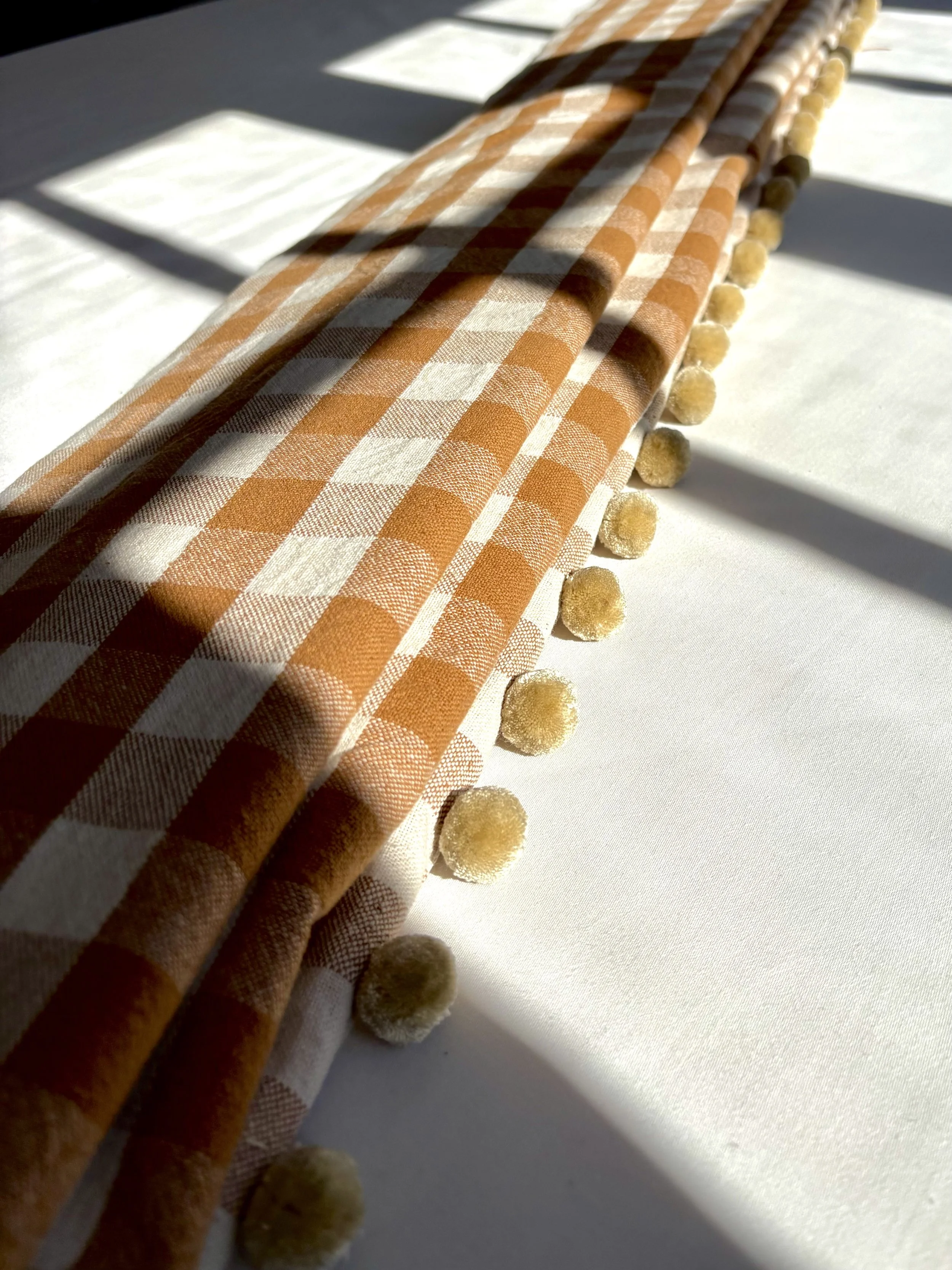 Close-up of orange and white checkered fabric with small yellow pom-poms along the edge, cast with shadows on a white surface.